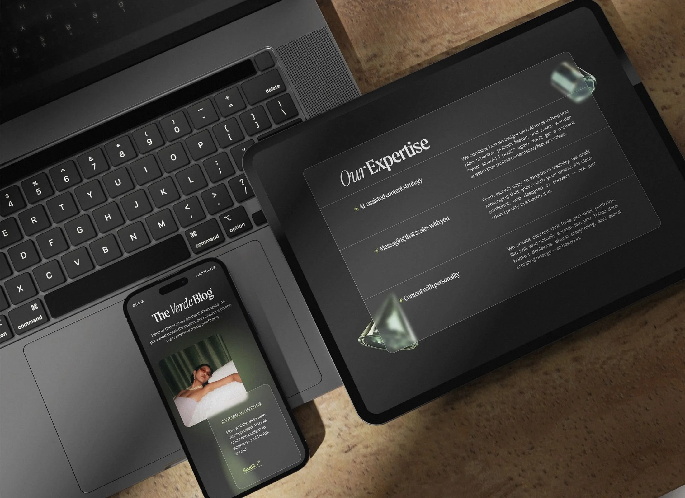 A laptop and a tablet on a wooden surface. The laptop screen shows a black background with text. The tablet displays a digital article titled 'Our Expertise' with bullet points on content strategy, messaging, and content with personality. A smartphone shows a blog post titled 'The Verde Blog' with an image of a woman lying on a bed and some text.