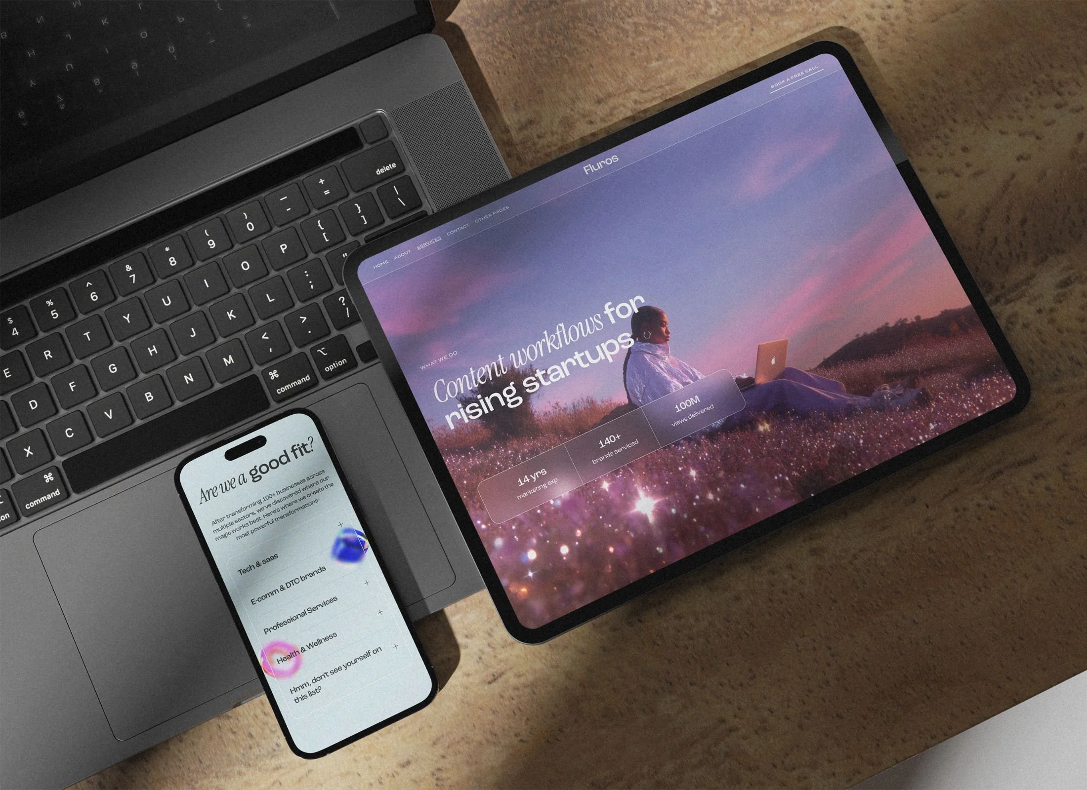 A laptop and a tablet on a wooden desk, with a smartphone resting on the laptop's touchpad. The laptop has a black keyboard and the tablet displays a website about content workflows for rising startups. The smartphone shows a list of categories like tech, SaaS, e-commerce, and health & wellness.