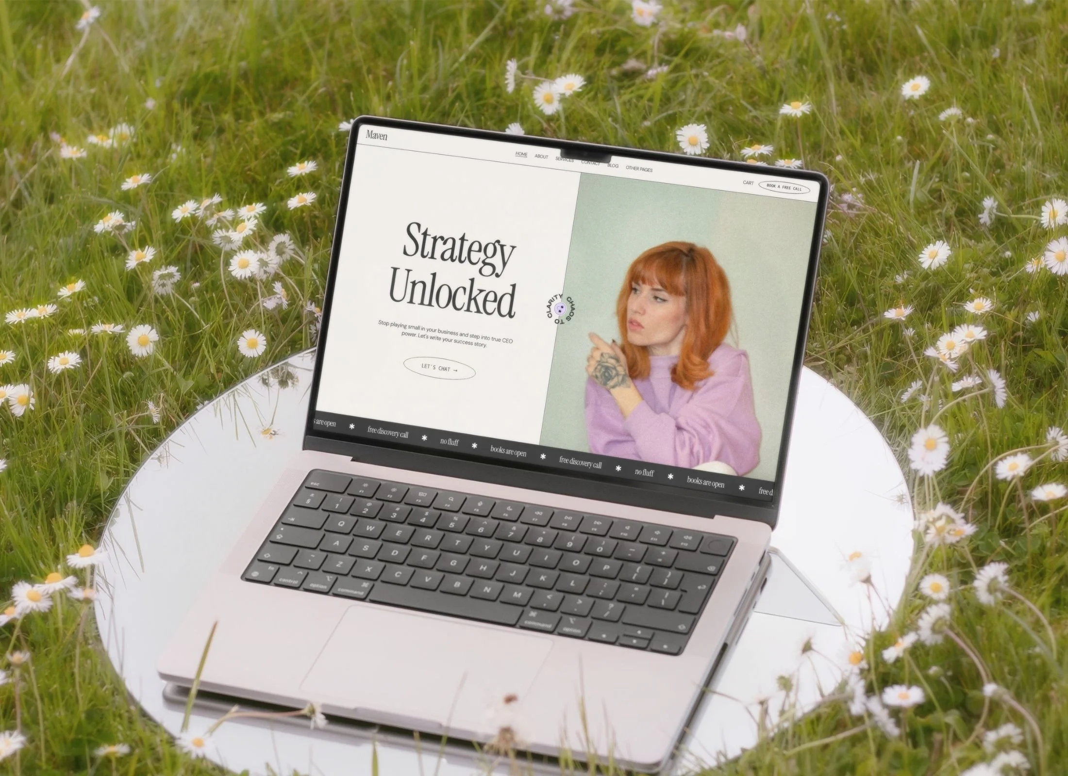A laptop on a circular mirror, set on grass with small white daisies. The laptop screen displays a website with the text "Strategy Unlocked" and an image of a woman with red hair, wearing a lavender top, pointing with a thoughtful expression.