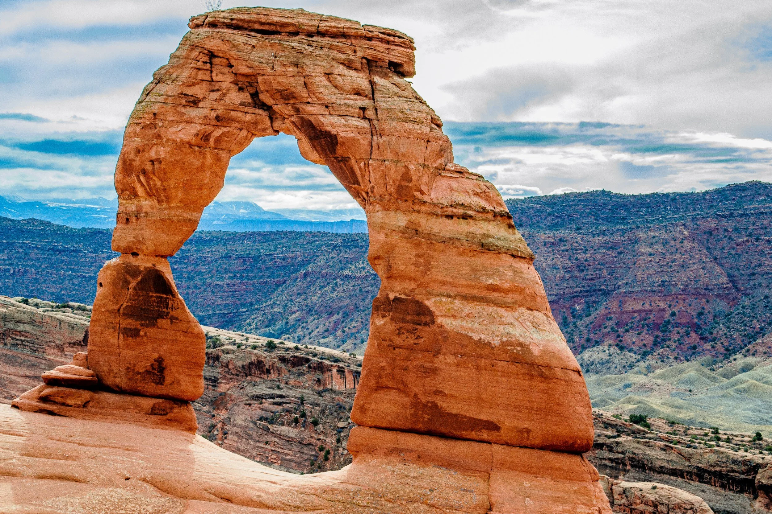 A natural rock formation known as Delicate Arch in Arches National Park, Utah, consisting of a large, freestanding arch-shaped sandstone rock set against a mountainous landscape with a partly cloudy sky.