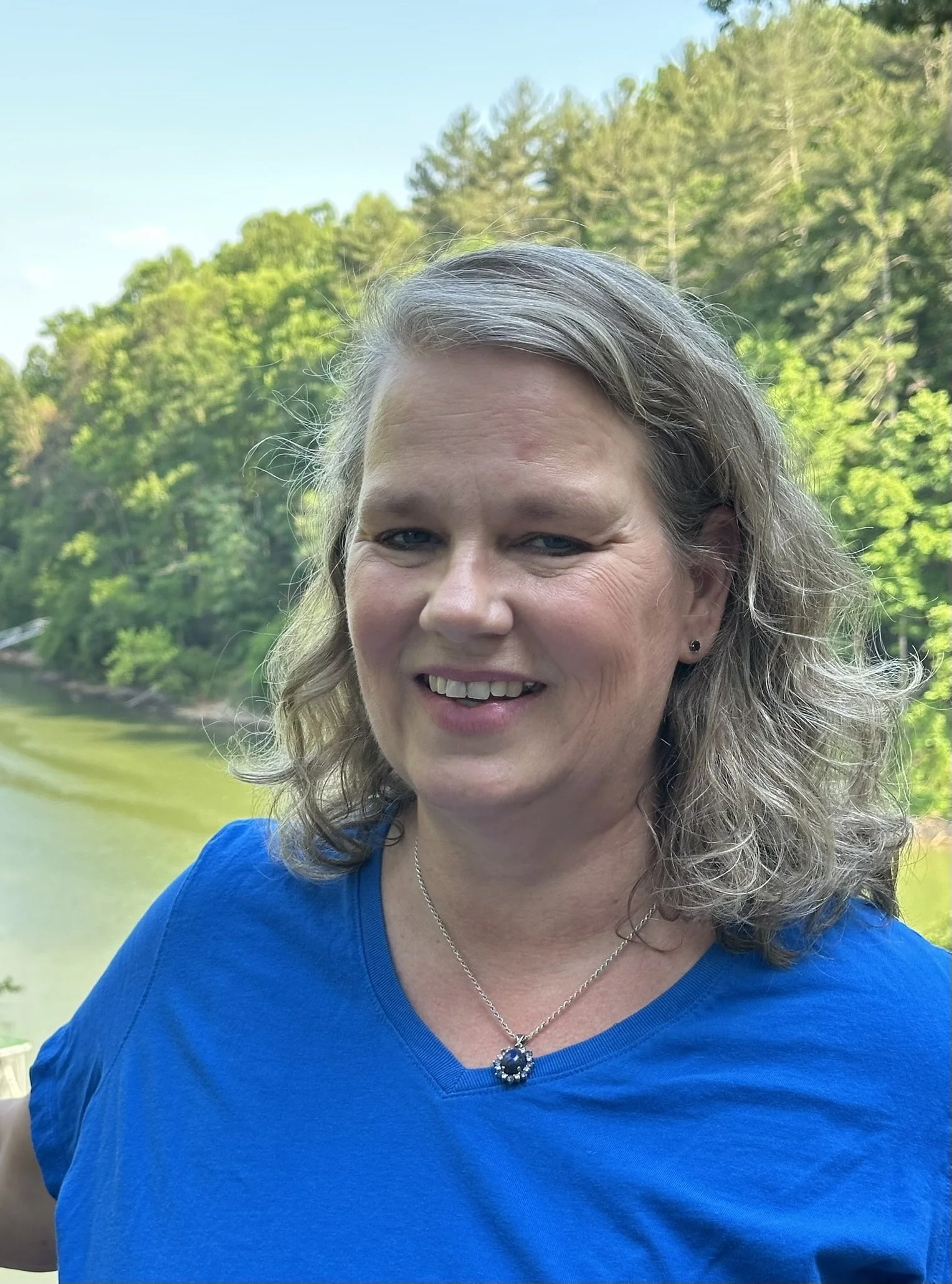 Close-up of a smiling woman with gray hair in a ponytail, wearing a blue shirt, standing outdoors with a background of trees and a body of water.