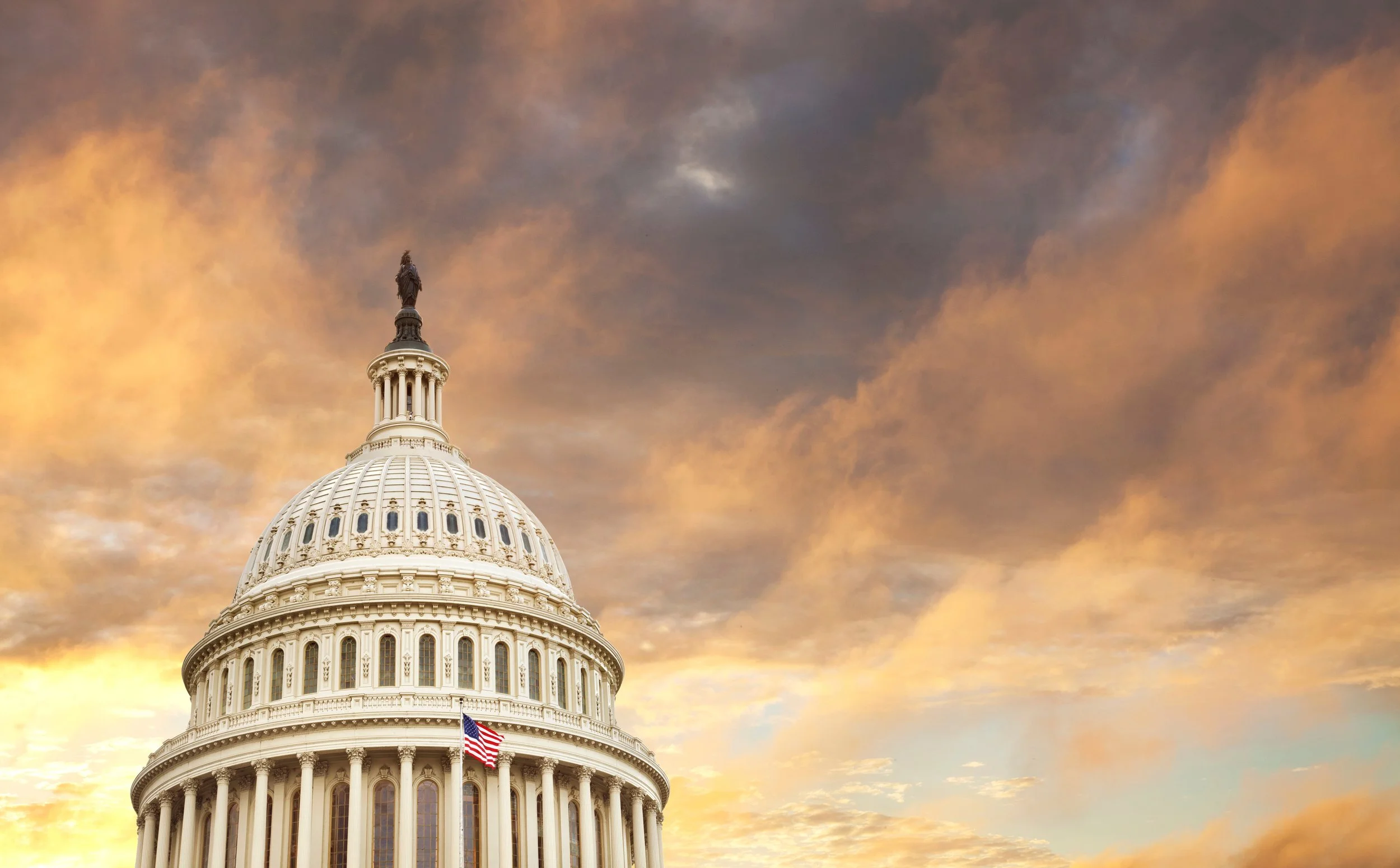 The Capitol building with a prominent dome, flag, and statue, set against a dramatic, cloudy sunset sky.