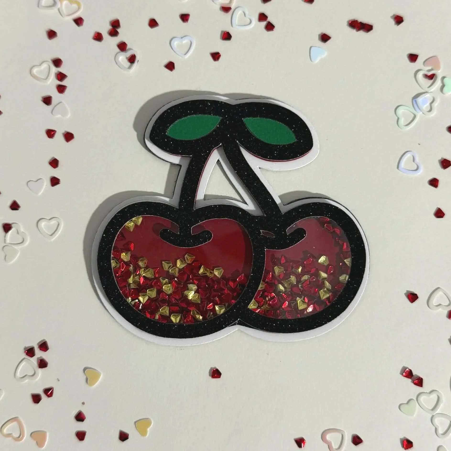 A magnet of two cherries with green leaves, outlined in black with glitter, and filled with red and gold heart-shaped confetti, surrounded by scattered red, white, and pastel heart-shaped confetti on a white background.
