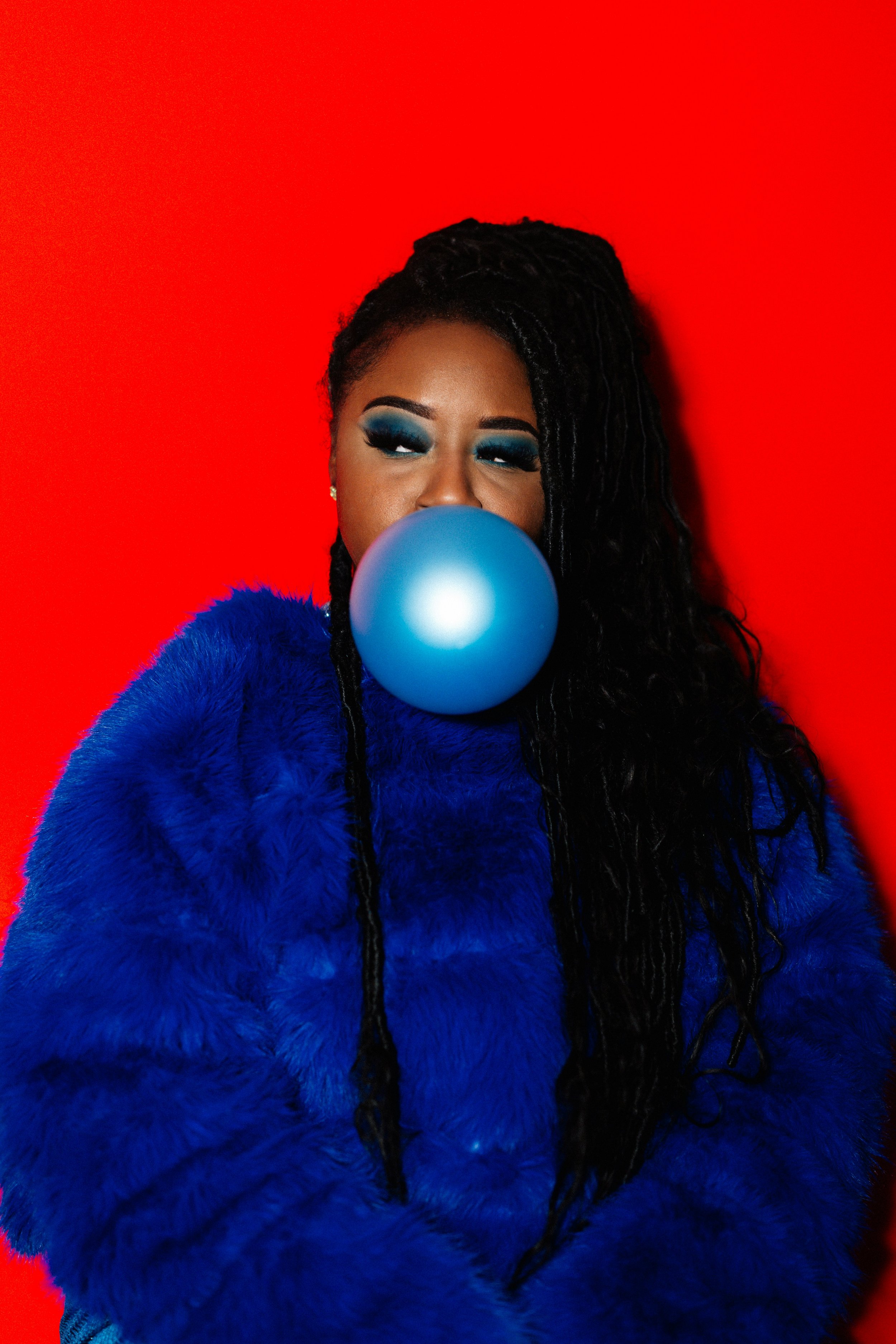 A woman with long black dreadlocks, dressed in a bright blue faux fur coat, blowing a large blue bubblegum bubble, standing against a vibrant red background.