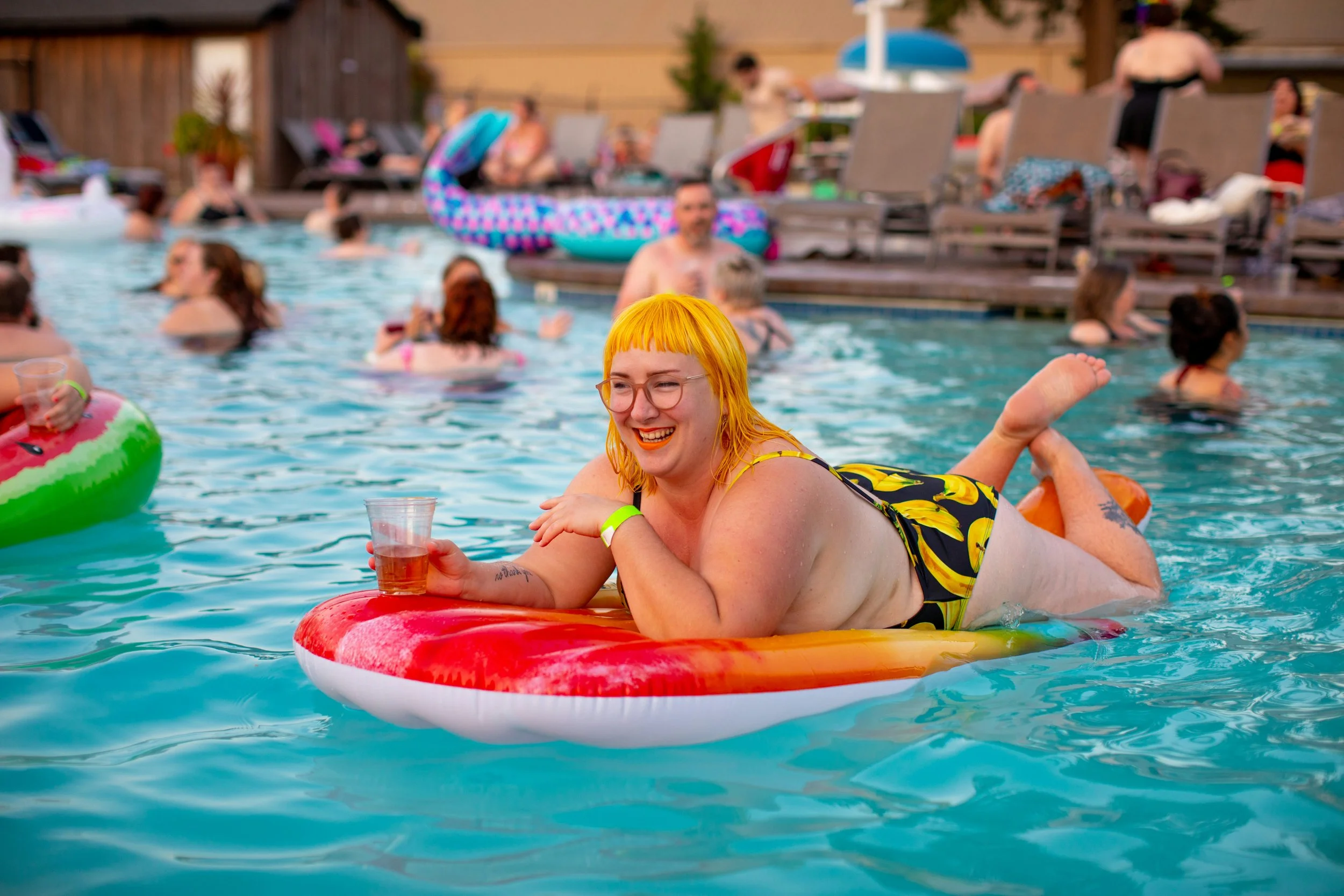 A woman with yellow hair and glasses relaxing on a float in a pool, holding a drink and smiling.