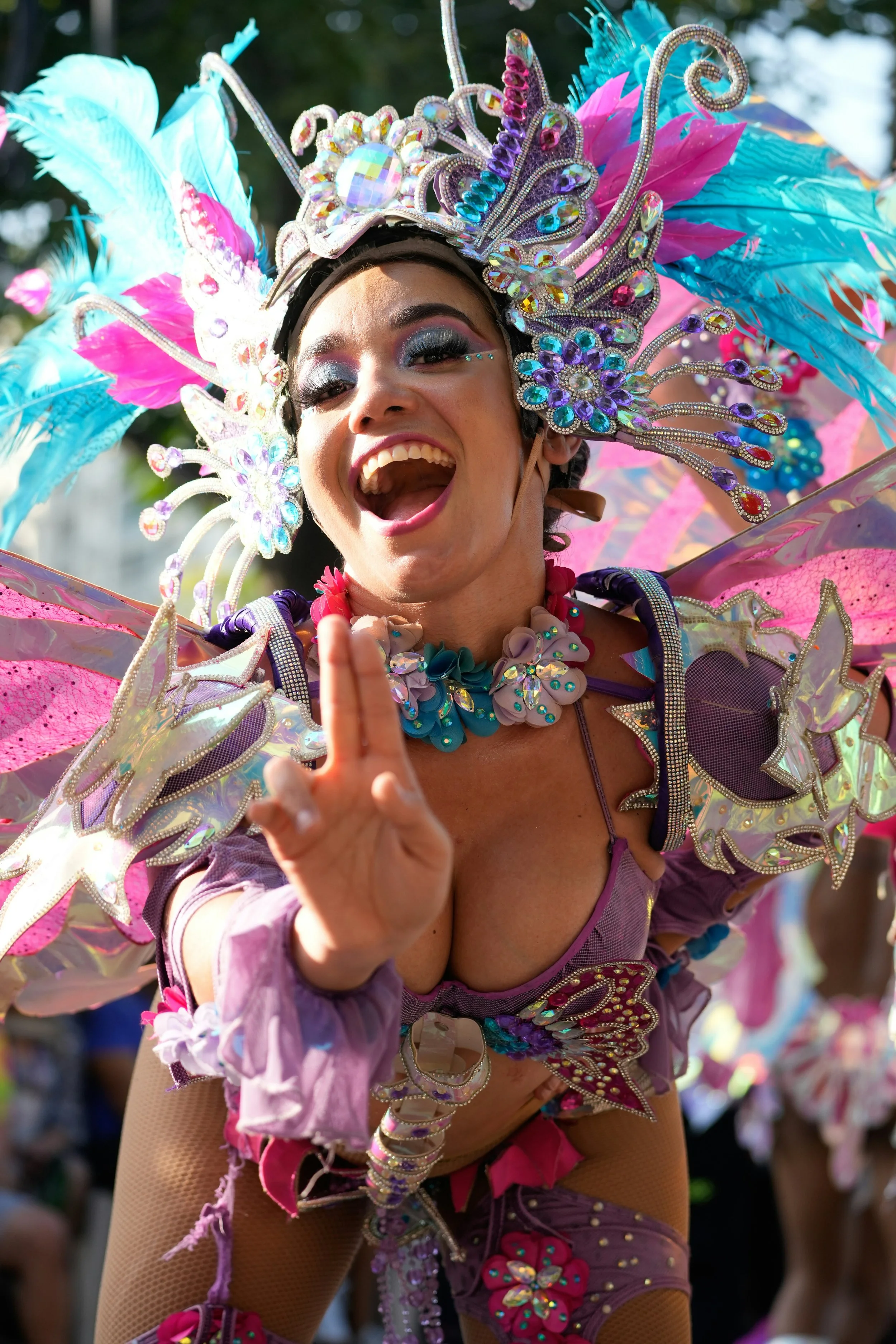 A woman in a colorful, elaborate costume celebrating at a carnival or parade, smiling and making a peace sign.
