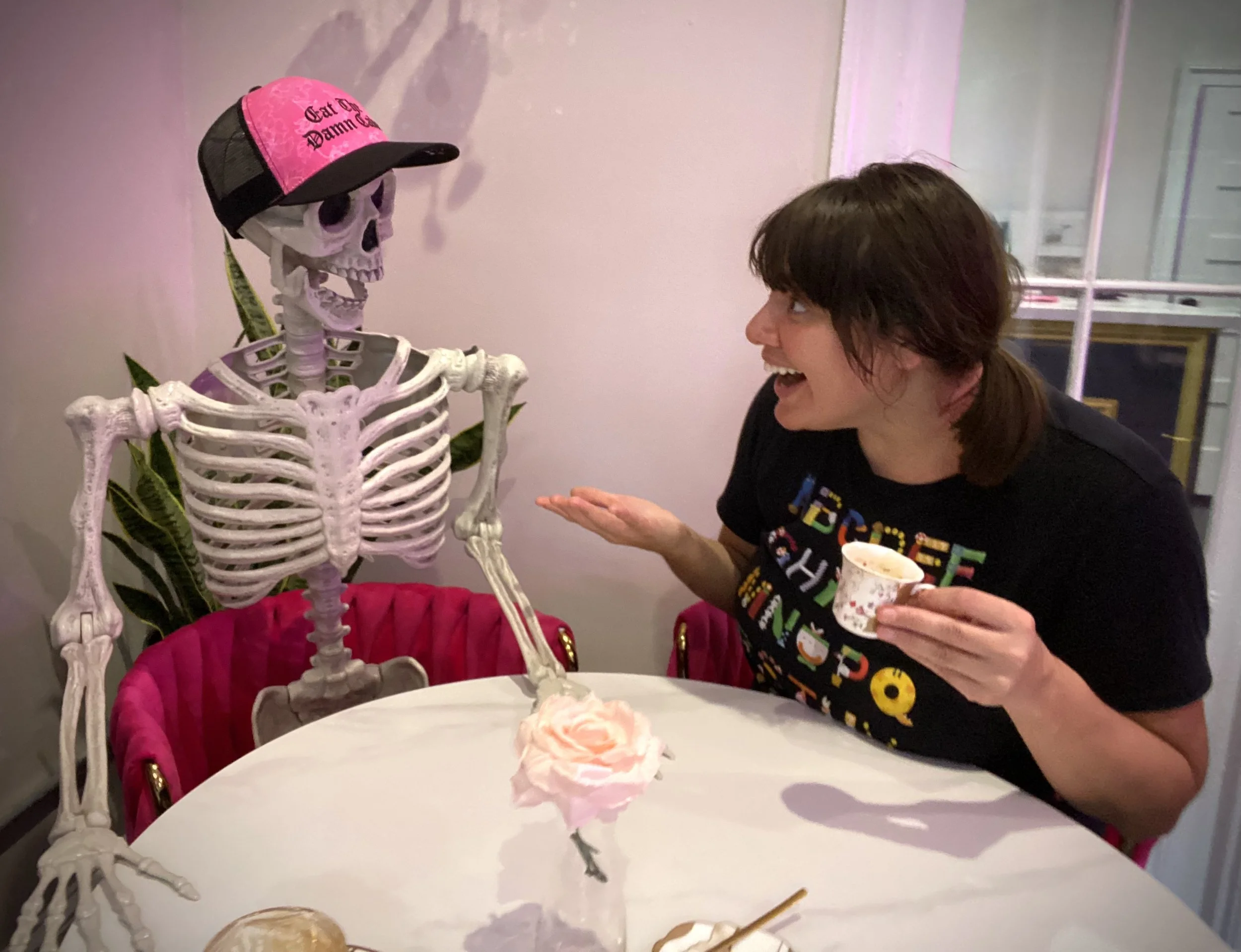 A woman with short brown hair and a black shirt is sitting at a table, smiling and talking to a skeleton wearing a pink and black cap. The woman is holding a small cup and gesturing with her hand. There is a pink flower on the table and some food ite