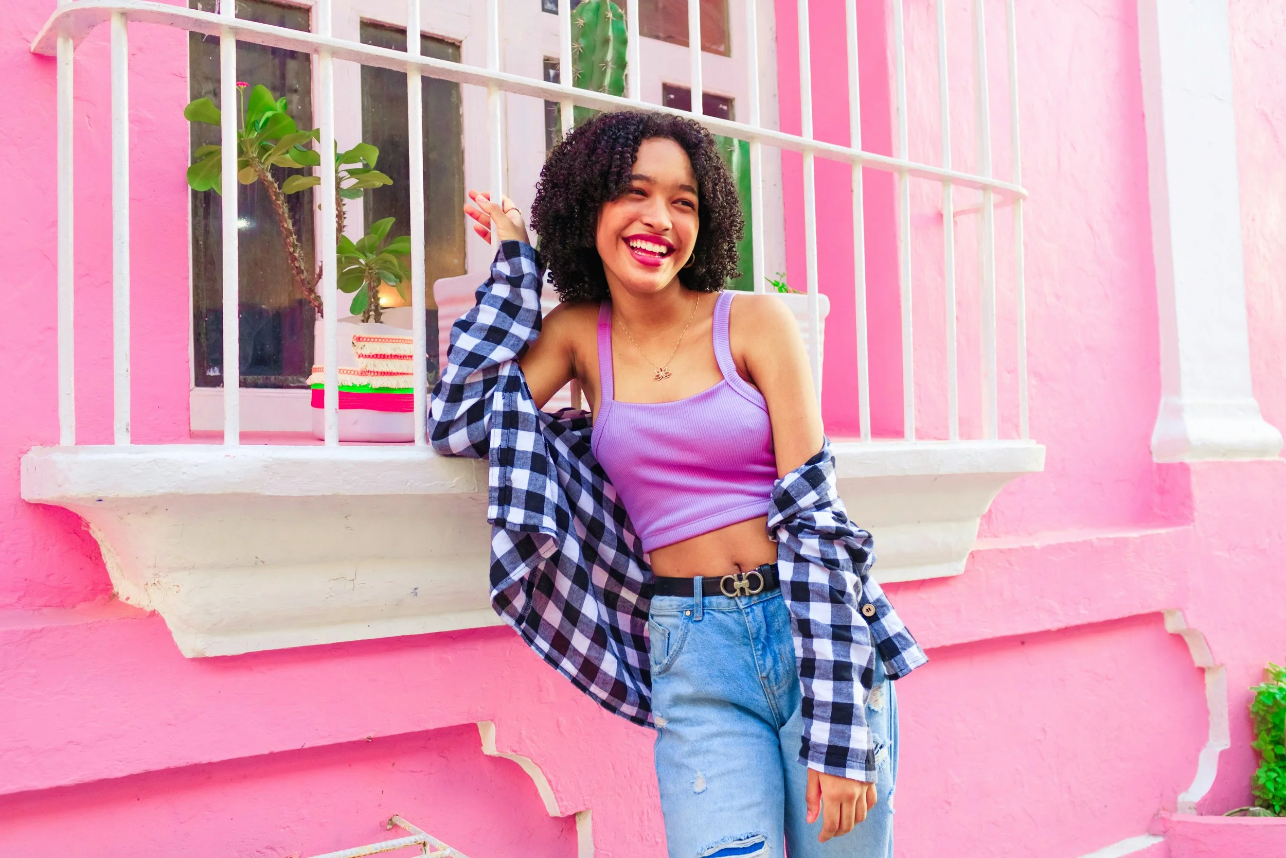 A smiling woman with curly hair wearing a purple crop top, ripped jeans, and a checkered shirt draped over her shoulders, standing in front of a pink wall with a window and green plants.