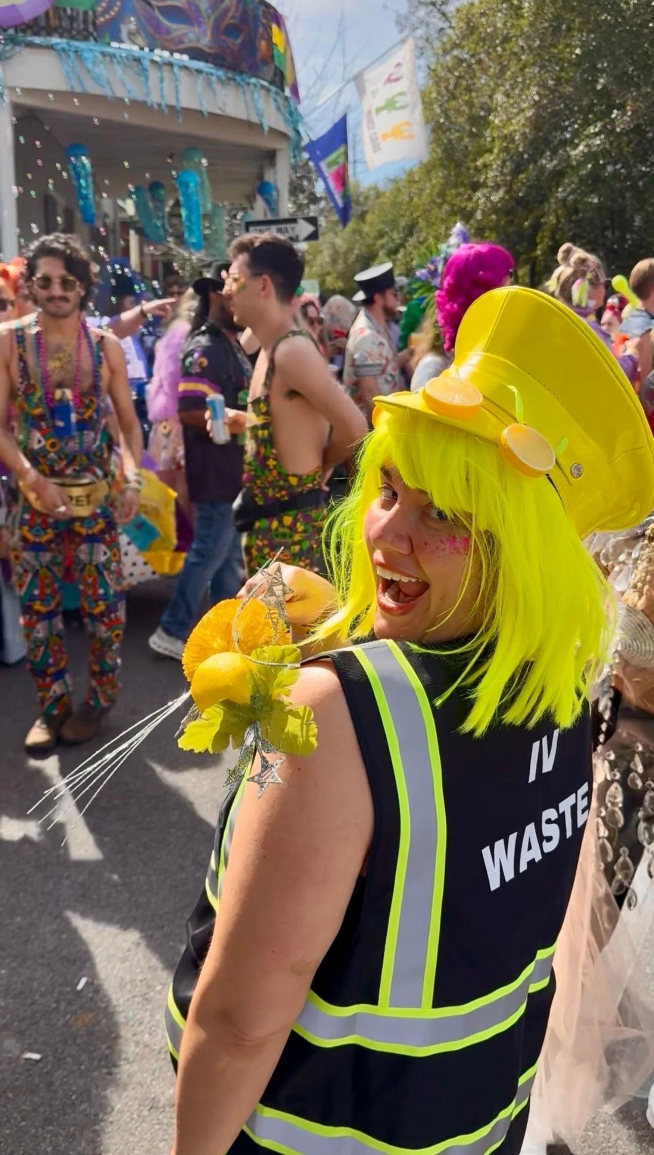 A woman with bright yellow hair, wearing a yellow painted helmet with lemon decorations, a black vest with reflective stripes that says 'IN WASTE,' at a lively outdoor parade or festival with people in colorful costumes in the background.
