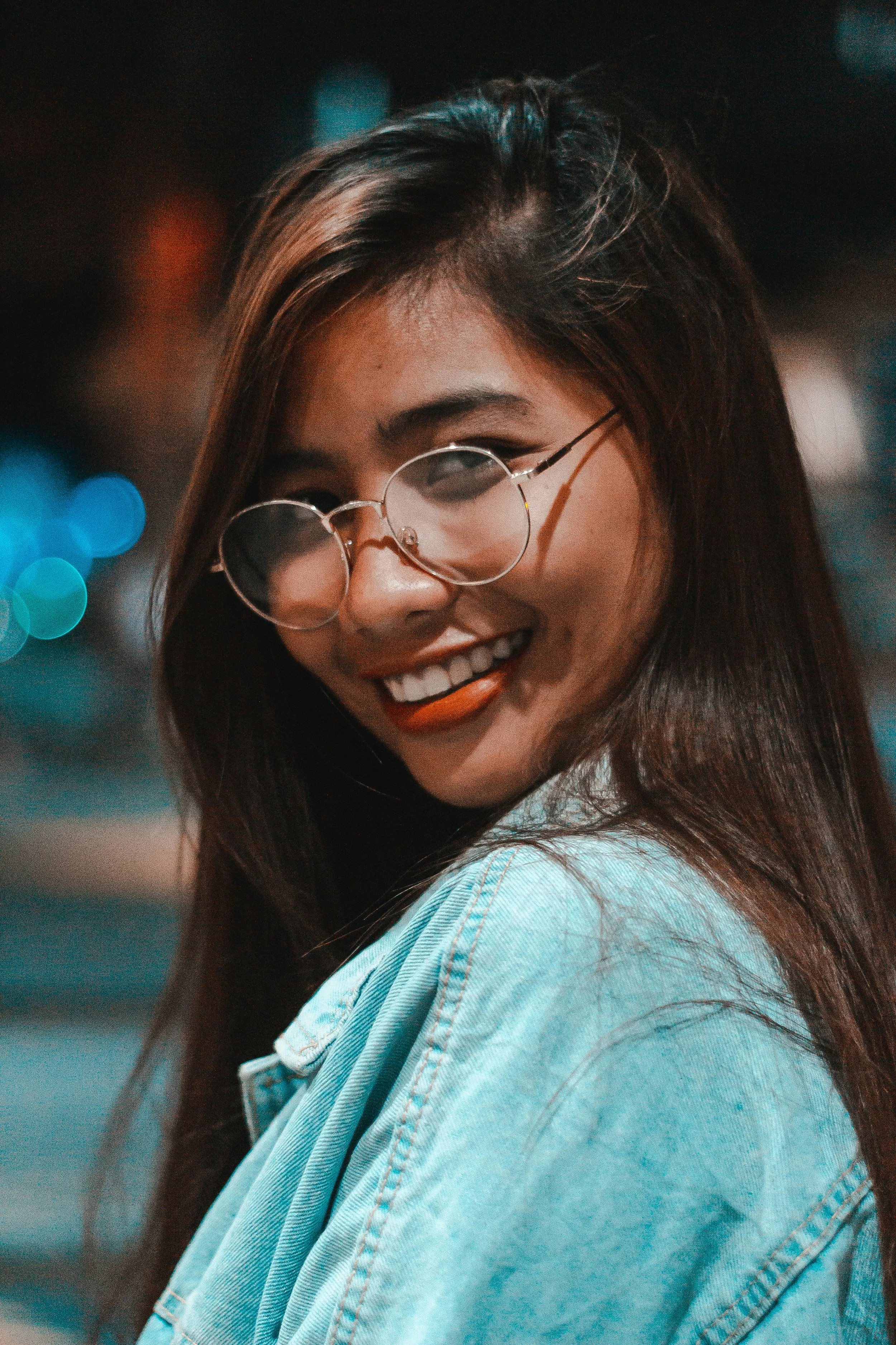 A young woman with long dark hair, wearing glasses and a light blue denim jacket, smiling at night with blurred city lights in the background.