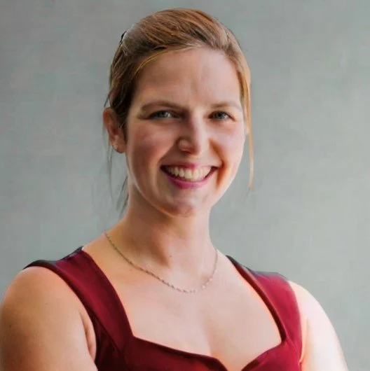 A smiling woman with reddish-brown hair tied back, wearing a navy blue sleeveless top and a delicate necklace, standing in front of a modern building with horizontal metallic panels.