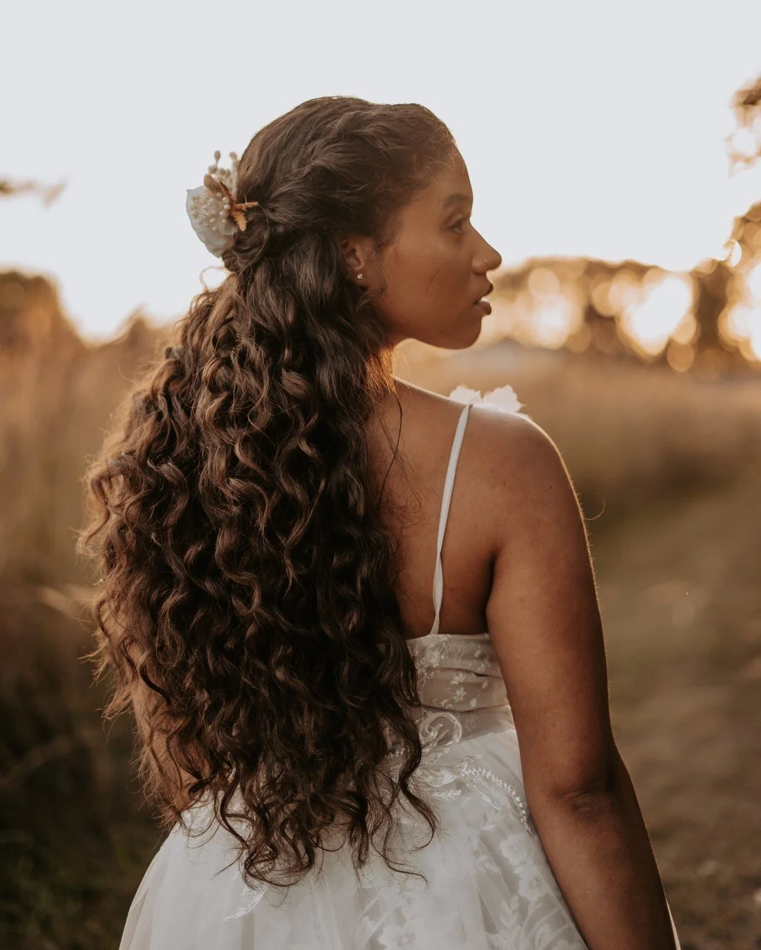 A woman with long, curly dark hair and a flower hair accessory, wearing a white dress with spaghetti straps, stands outdoors during sunset in a natural setting.