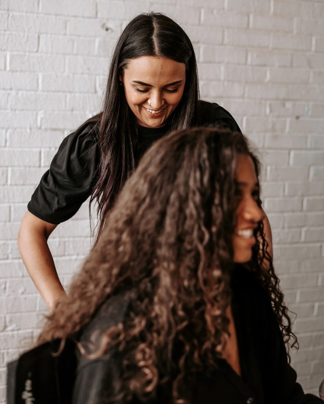 Two women smiling, one with straight black hair and the other with curly brown hair, sitting against a white brick wall.