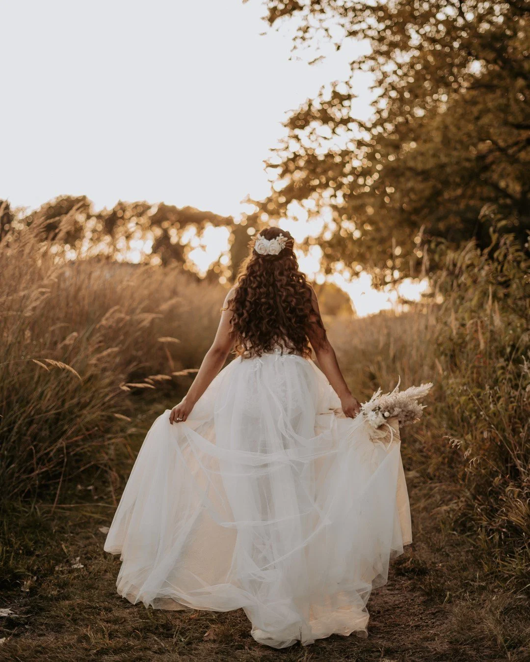 A woman with long, curly hair and a white flower crown walking through a field of tall grass during sunset, wearing a white dress and holding a bouquet.