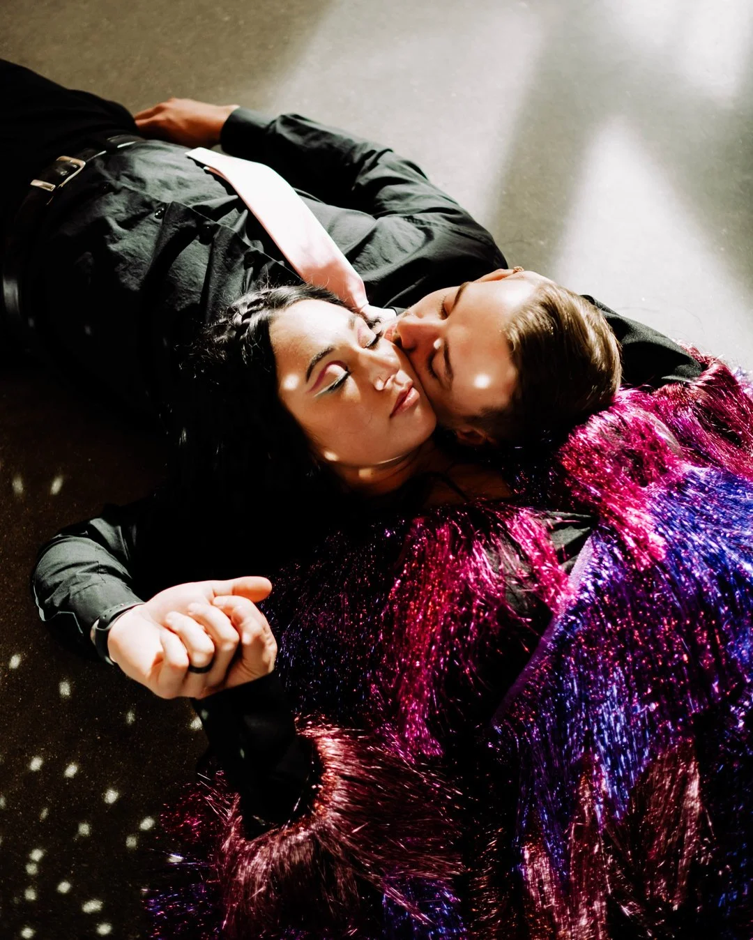 Two people are lying on the floor, embracing. One is wearing a black outfit with a pink tie, and the other is dressed in a colorful, sparkly garment. They are close, with their faces touching, suggesting an intimate or emotional moment.