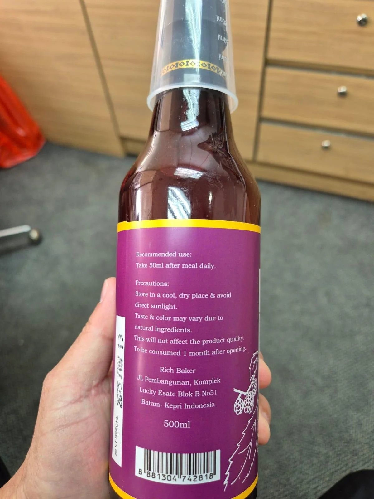 A person holding a 500ml bottle with a purple label. The label includes instructions for use, precautions, and manufacturing details, with a barcode at the bottom. The bottle appears to contain a beverage or liquid product, with part of a plastic cap