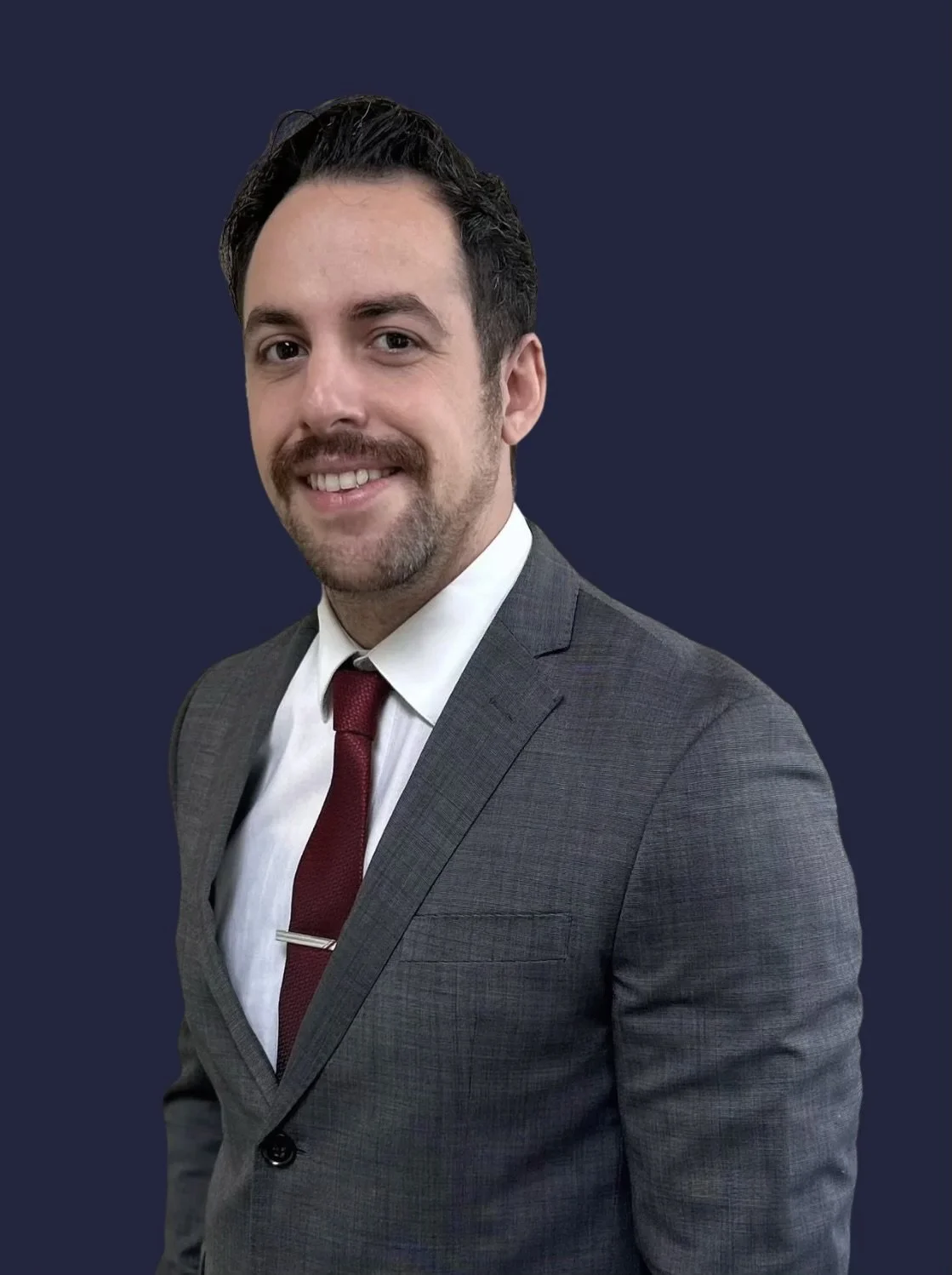A man wearing a gray suit, white shirt, and red tie, smiling at the camera, with a transparent background.