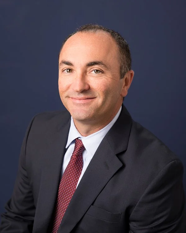 A middle-aged woman with short blonde hair, wearing a black blazer over a striped dress shirt, and a pearl necklace, smiling against a black background.