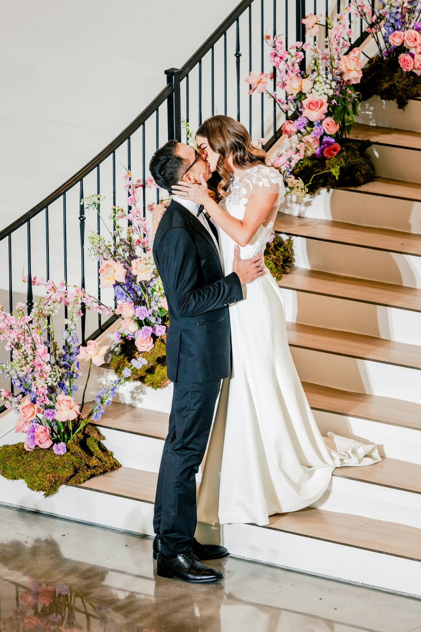 A newlywed couple sharing a kiss on staircase decorated with pink, purple, and white flowers.