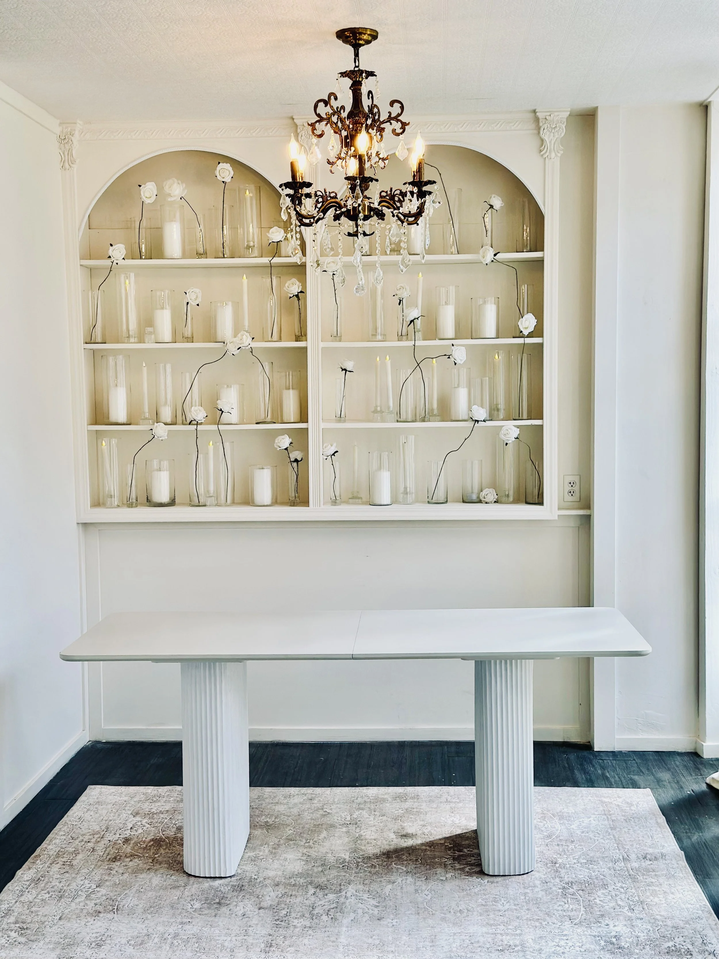 Modern white table with fluted legs in a room with a built-in white shelf filled with candles and white flowers in glass vases. A black chandelier with crystal accents hangs from the ceiling, and a beige area rug covers part of the dark hardwood floo