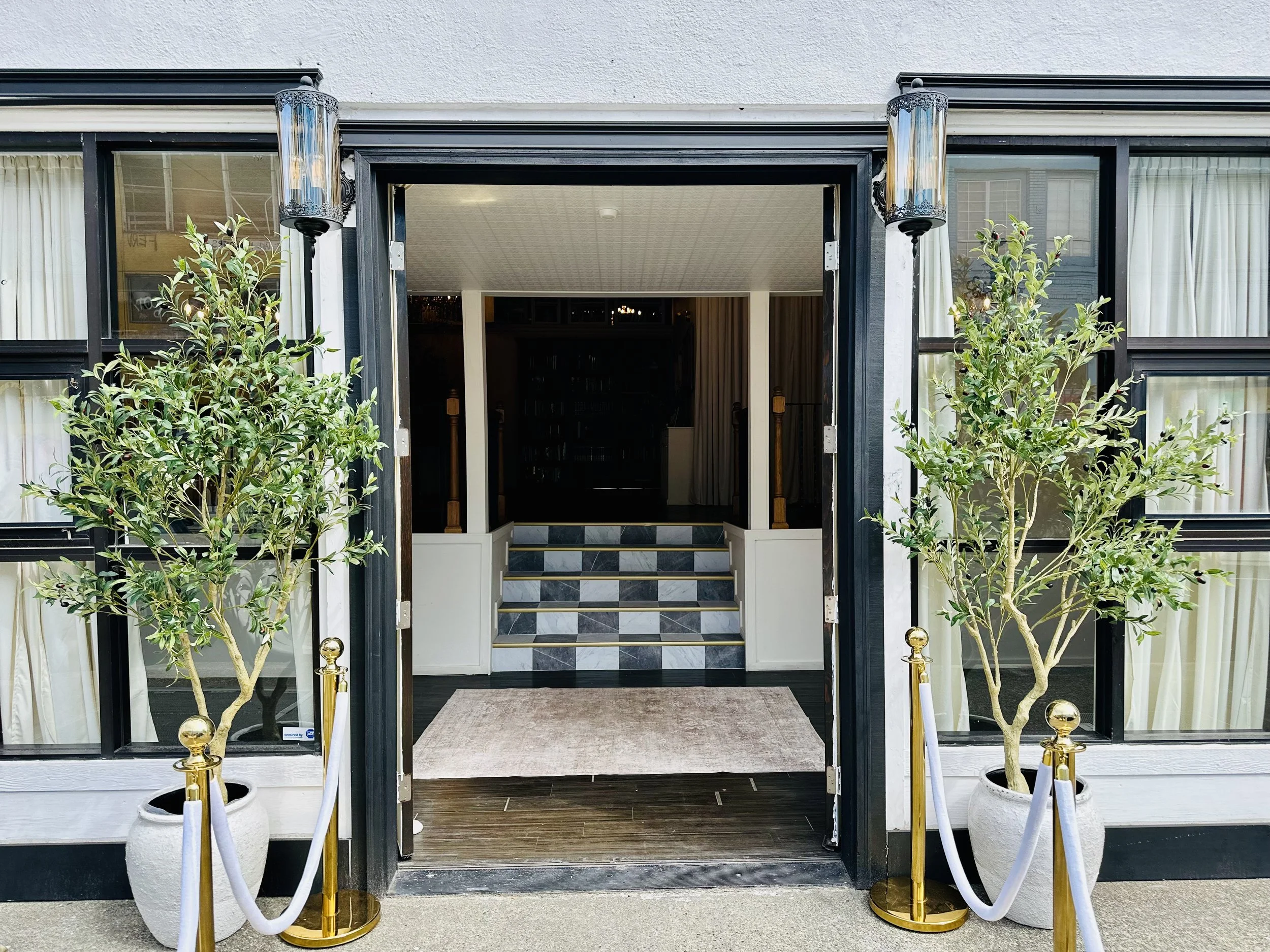 Front entrance of a building with double doors, two potted plants on either side, and decorative lights overhead.