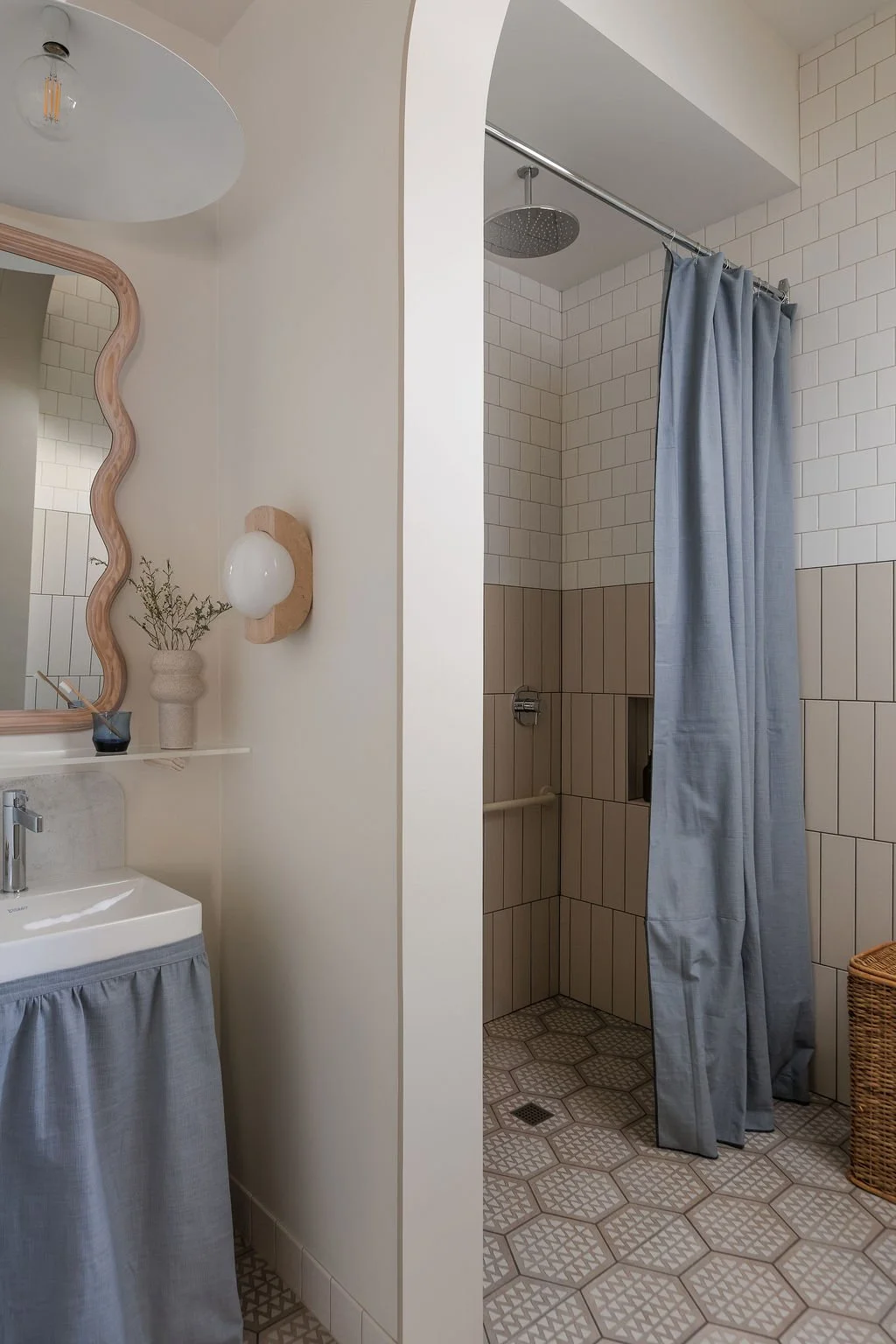 Washroom vanity with wall sconce and wavy mirror. Accessible shower with long blue shower curtain.
