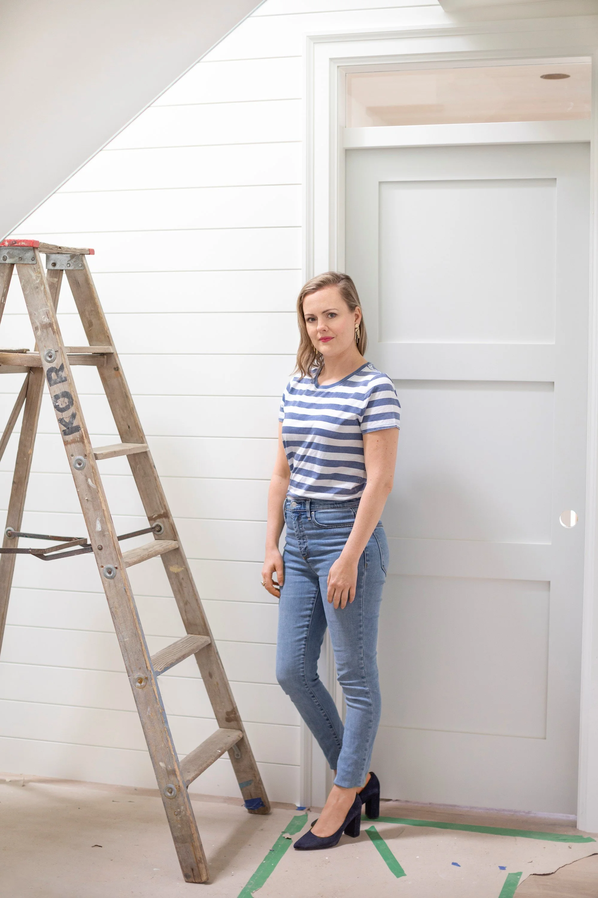 A woman with light brown hair wearing a blue and white striped t-shirt, blue jeans, and black high heels standing next to a wooden ladder in a room with white paneled walls and a white door.