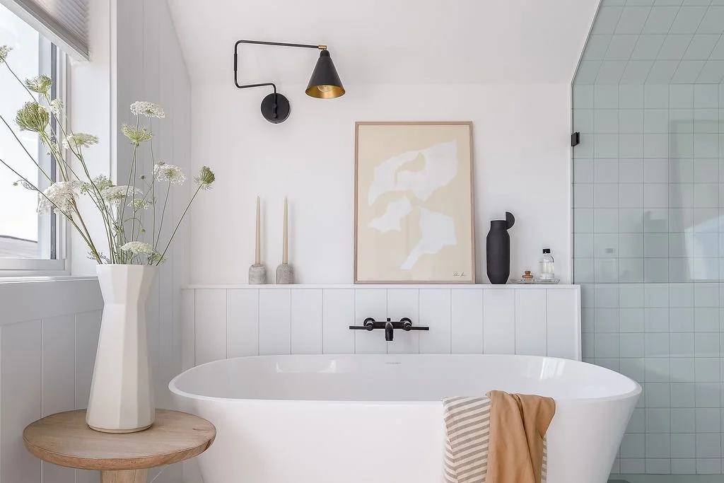 Soaker tub with art and decor on ledge above, flowers in vase on side table next to window.
