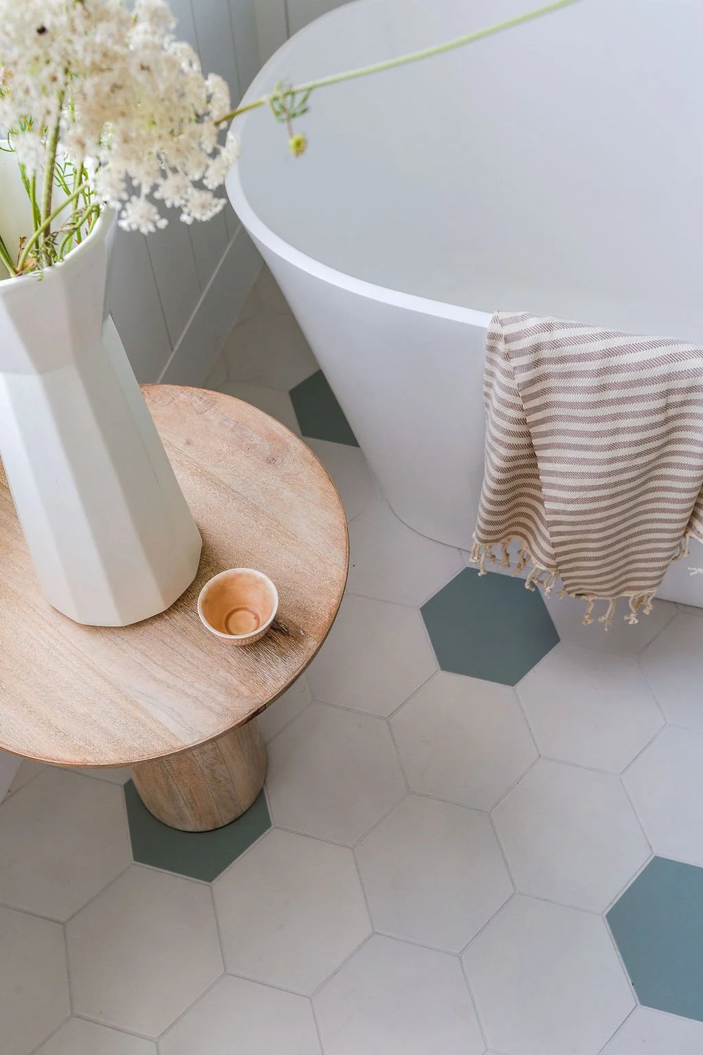 close-up of table on large white and green hexagon flooring, wood table with flowers in vase and striped towel draped over edge of tub.