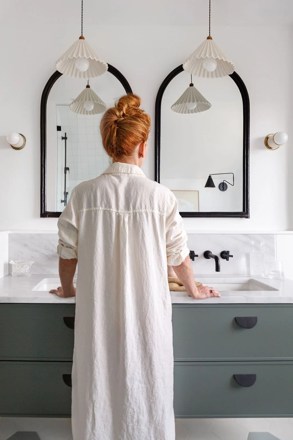 Woman with red hair wearing long linen shirt standing at green washroom vanity with pendant plighting and arched mirrors.