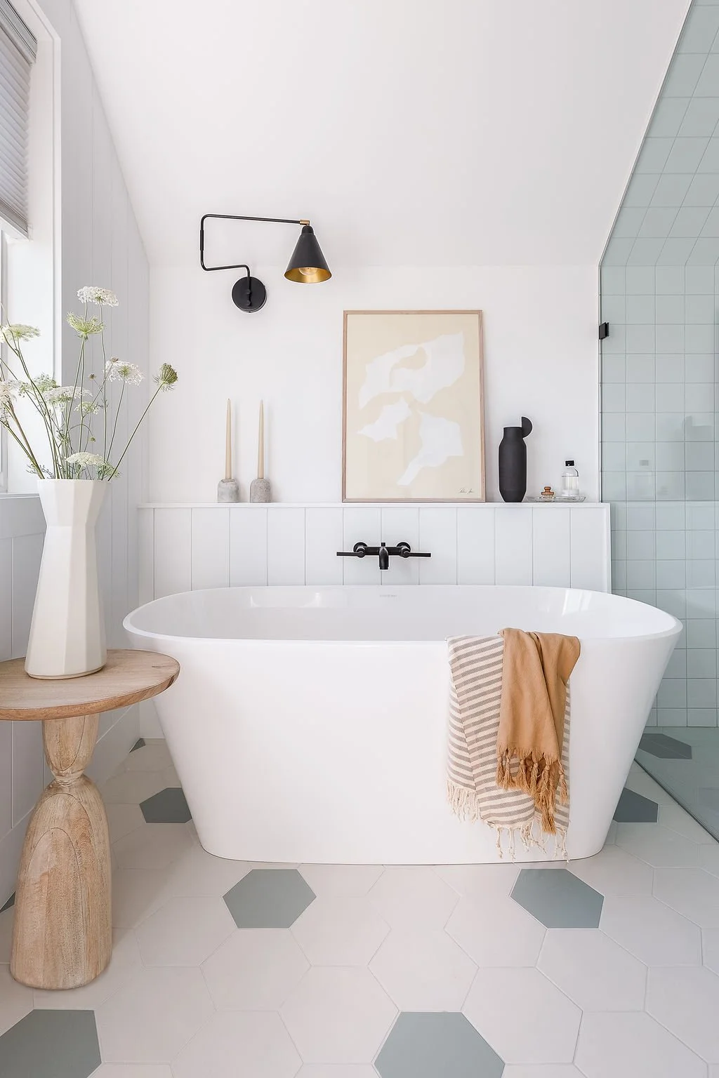 Soaker tub with art and decor on ledge above, flowers in vase on side table next to window.