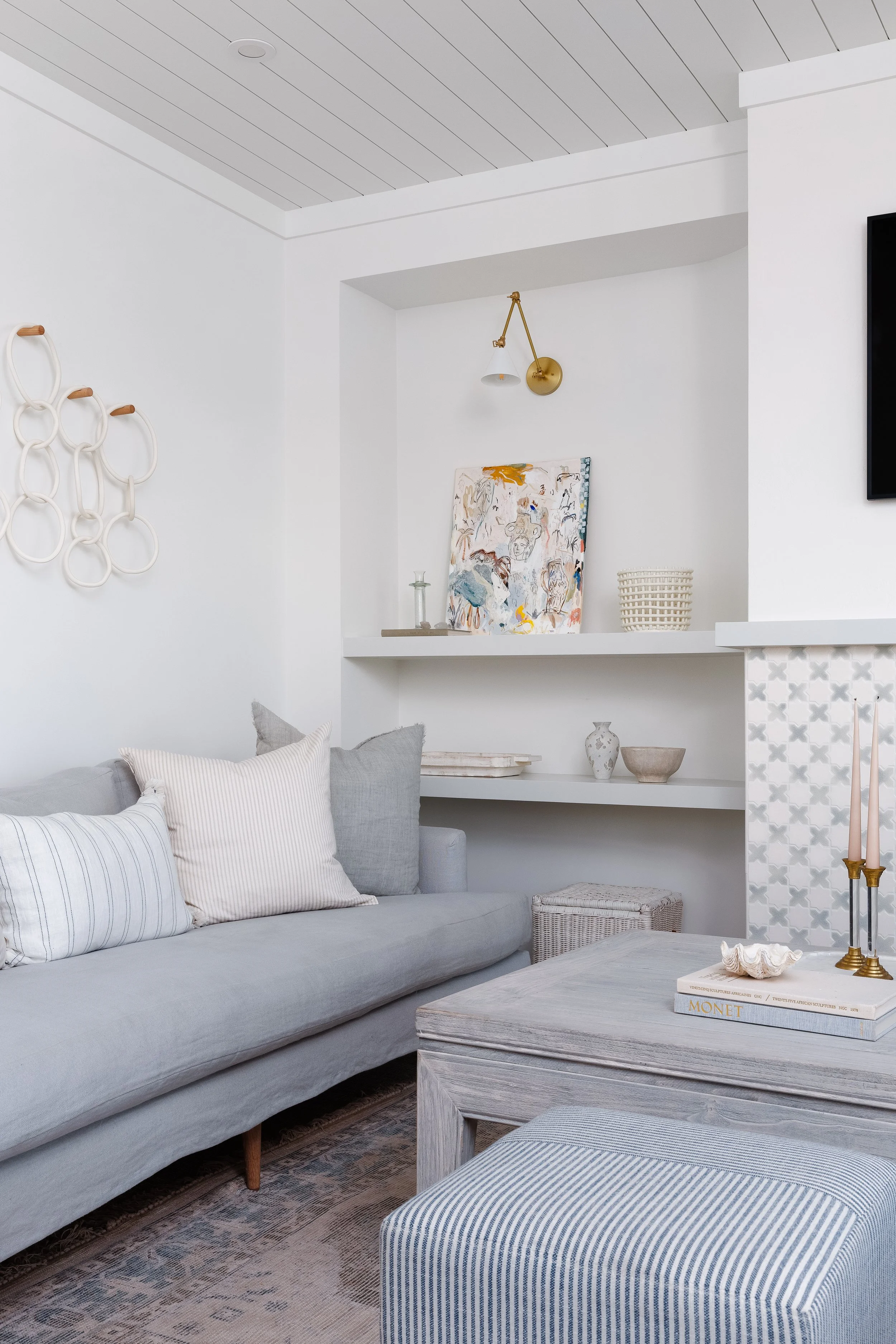 Fireplace with pretty white and blue mosaic tiles. Floating shelf next to fireplace with art and decor creatively displayed. Light blue linene sofa with push accent pillows, wood coffee table with candles. Striped ottoman.