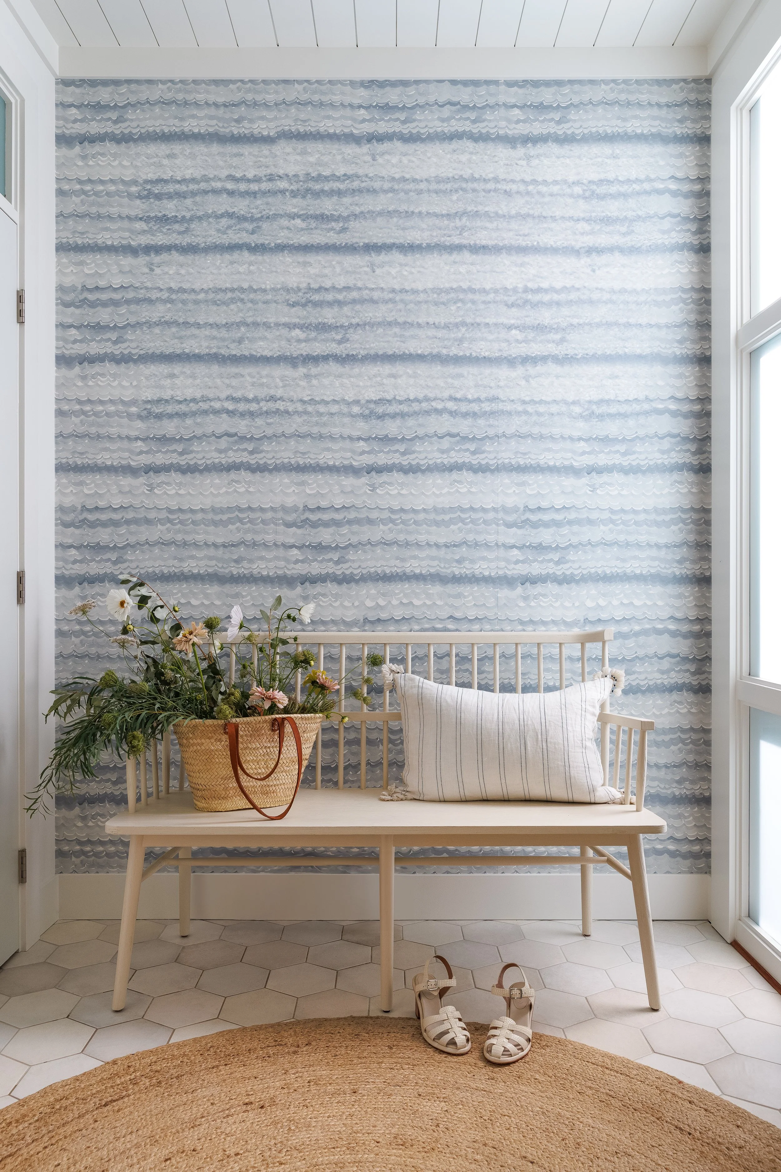Home entry with white spindle bench, striped toss cushion and basket with fresh flowers. Wall behind bench is wallpapered in small wave pattern wallpaper. 