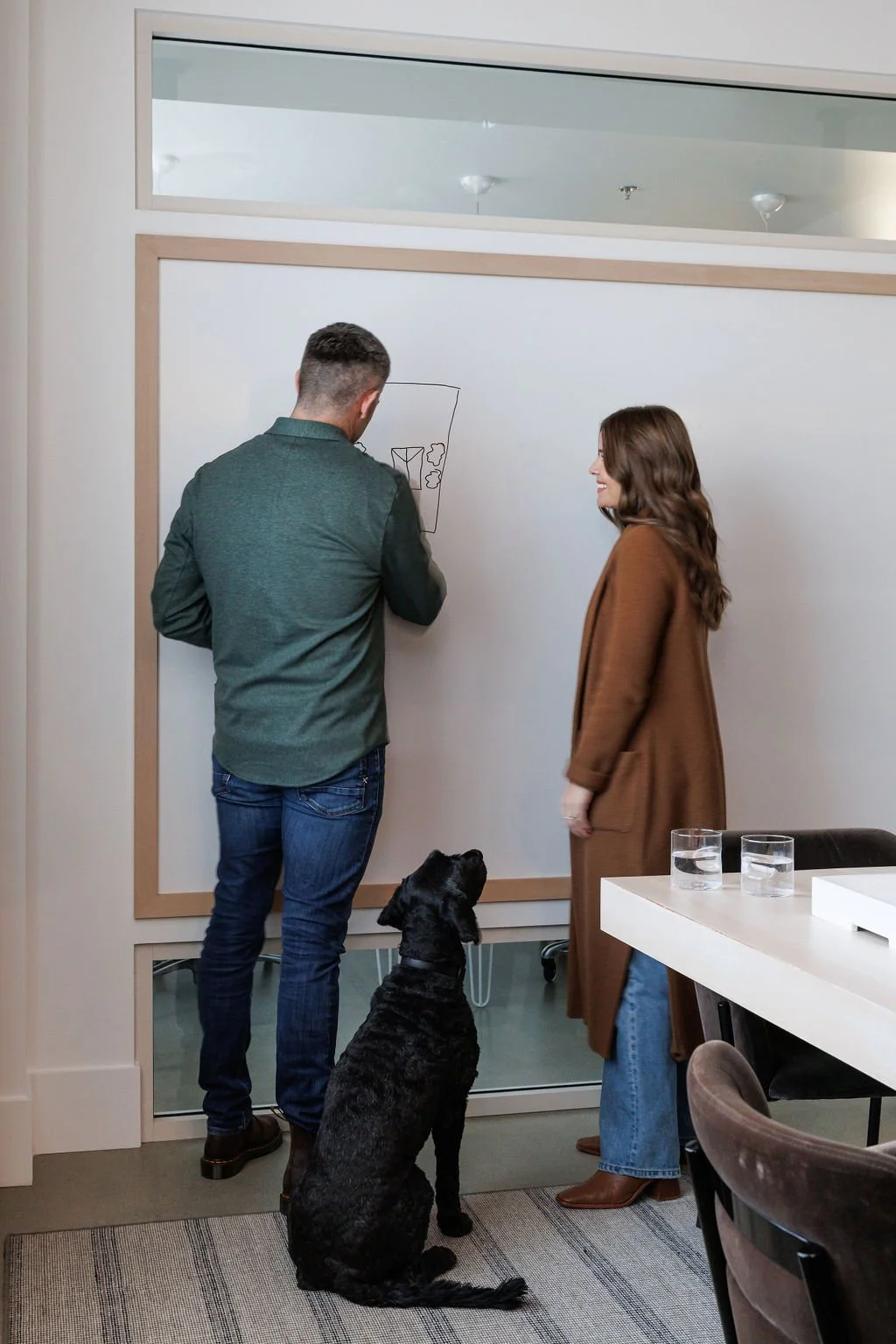Photo of man and woman drawing and discussing at white board. Black do is sitting next to man and woman.