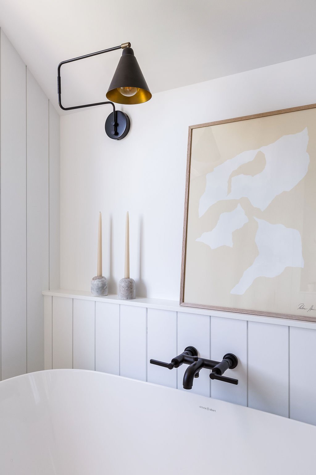 Pastel art and decor on ledge next to tub with black hardware and feature light.