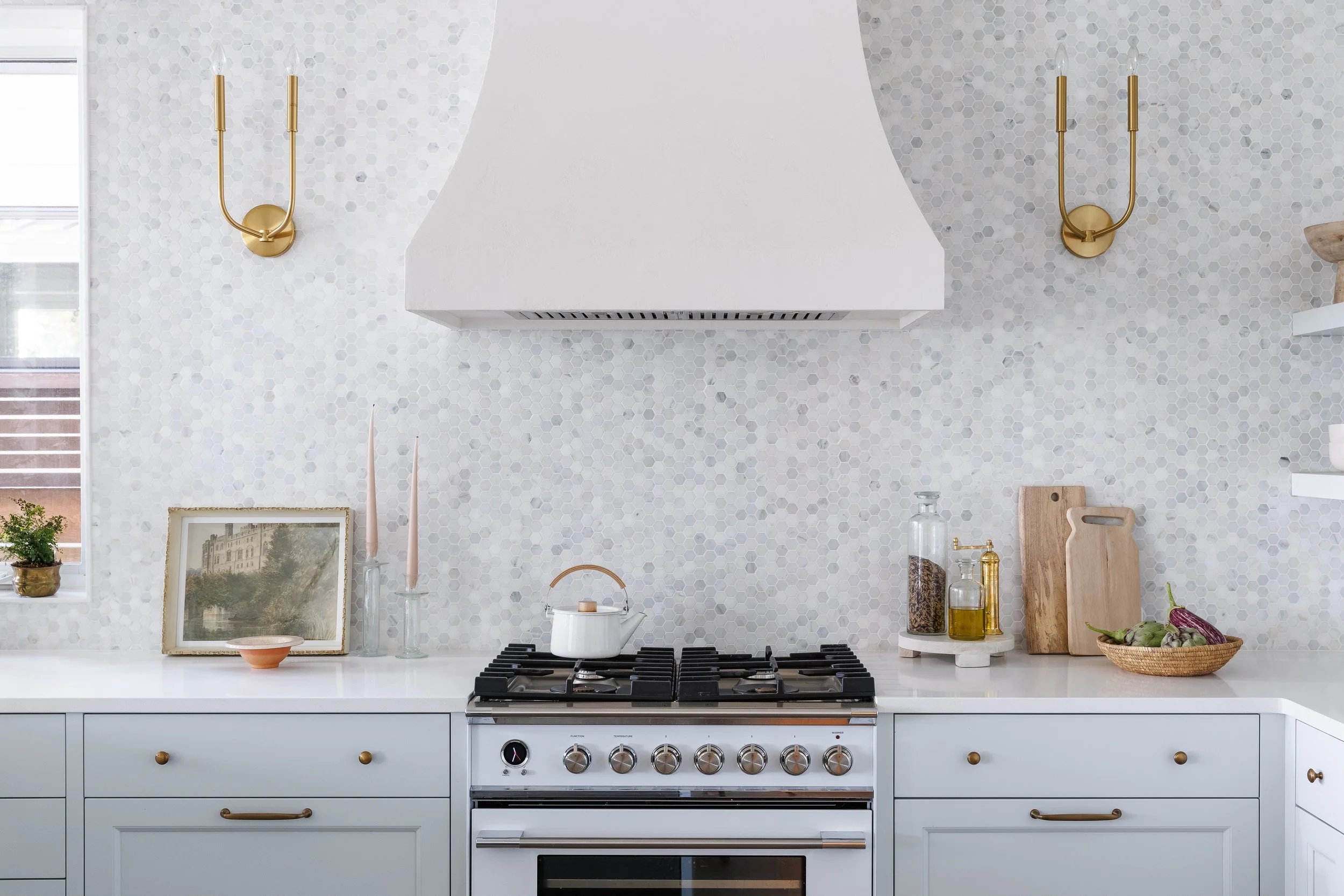 Close-up of light blue cabinetry with gold hardware, white range and curved plaster range hood. Counters styled with cutting boards, decanters with rice and oil, art and candle sticks.
