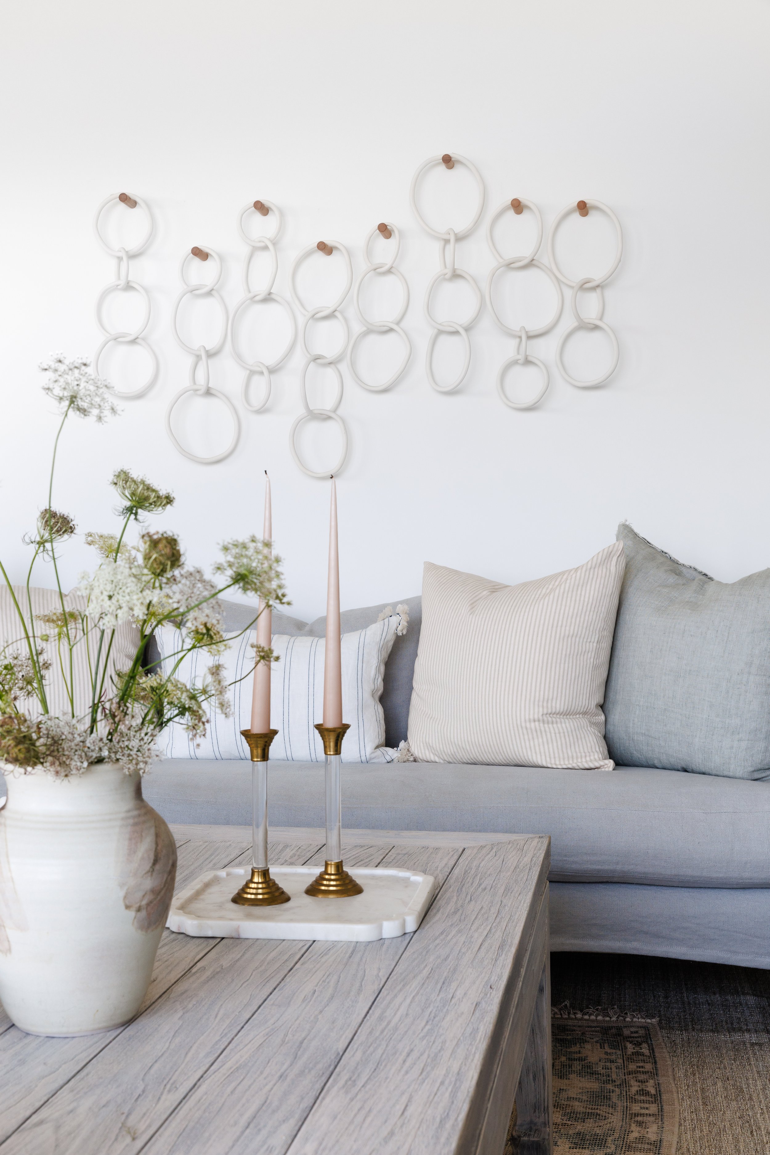 Coffee table with candle sticks and pottery vase with summer flowers. Light blue linen sofa with push accent toss cushions. Clay hoop artwork on wall above sofa..