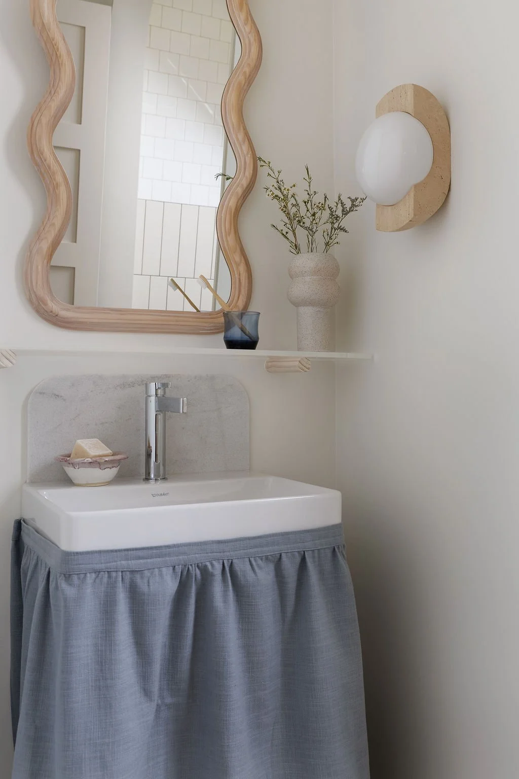 Washroom sink with curtain privacy panel, shelf above sink, wavy mirror and wall sconce.