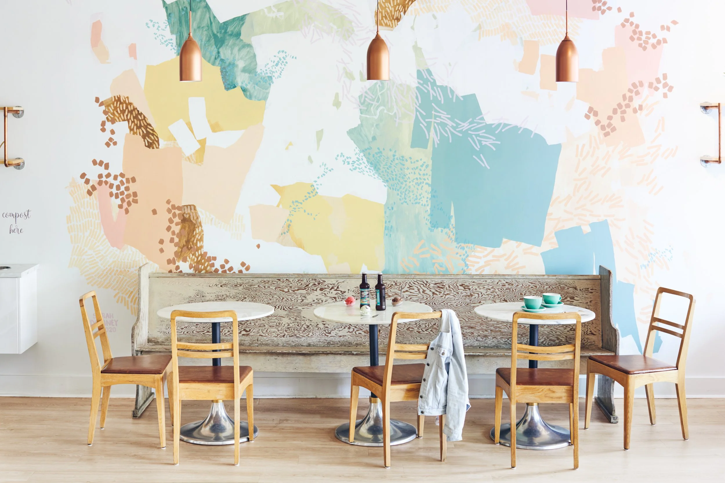 Rustic bench and bistro tables and chairs in gelato shop with artist mural on wall, gelato scoops on table and root beer.