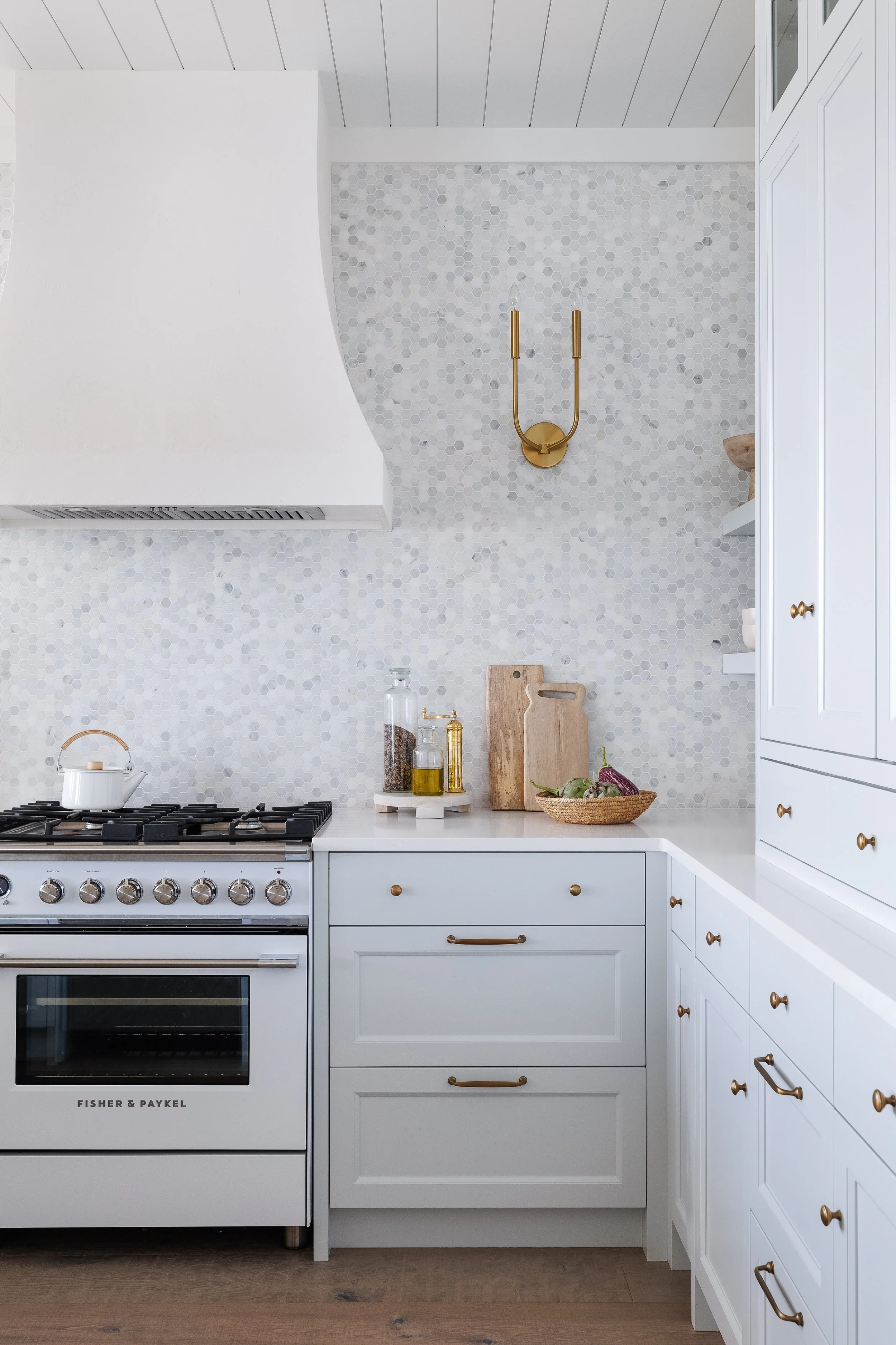 Corner of kitchen with marble hexagon mosaic light blue cabinetry with gold hardware, white range and curved plaster hood fan.