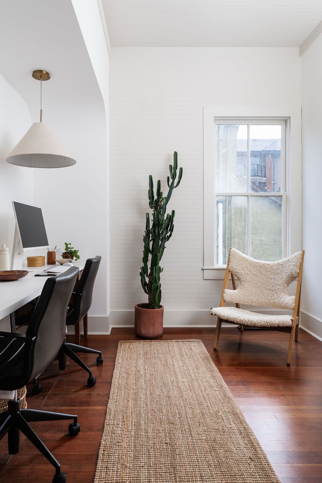 White office with black office chairs and white desk with arched builkhead and cone linen pendant above. Wool and wood chair next to window. 