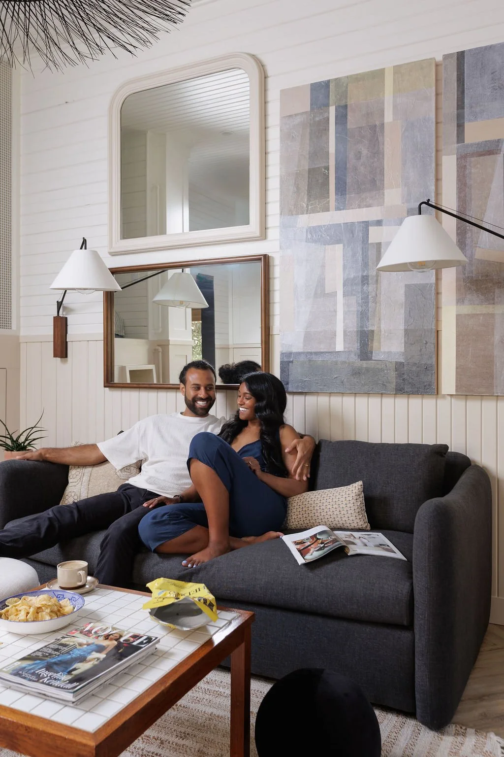 Rasila and Siya Herman snuggle on a charcoal sofa next to coffee table with magasines and snacks.