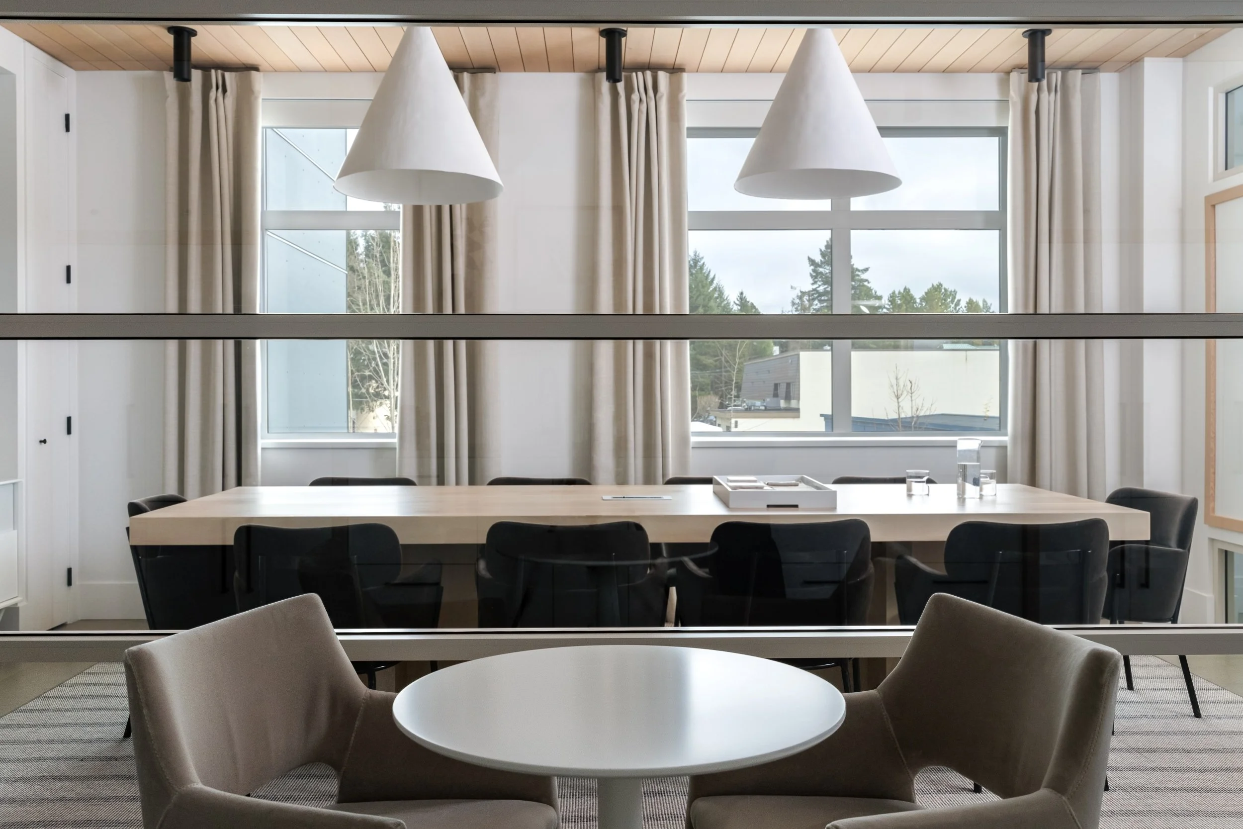 Looking through glass wall into boardroom and view outside. There is a wood boardroom table with brown velvet chairs and large white pendant lights over table.