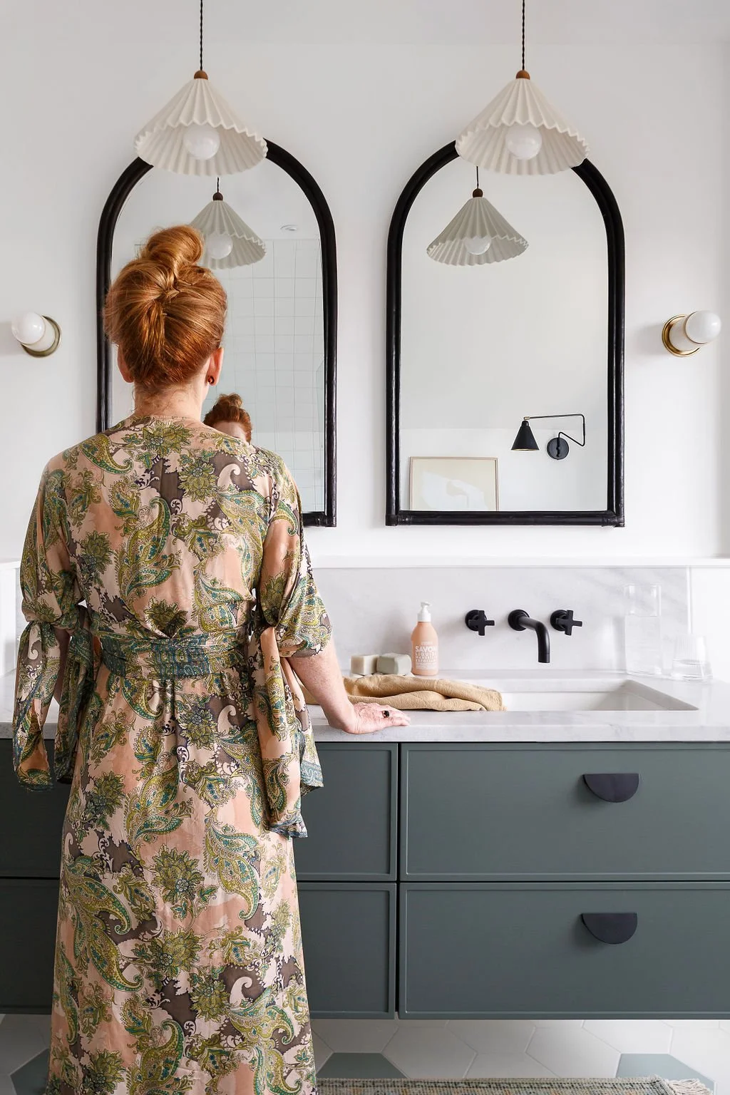 Woman with red hair wearing long green and peach paisley robe standing at green washroom vanity with pendant plighting and arched mirrors.