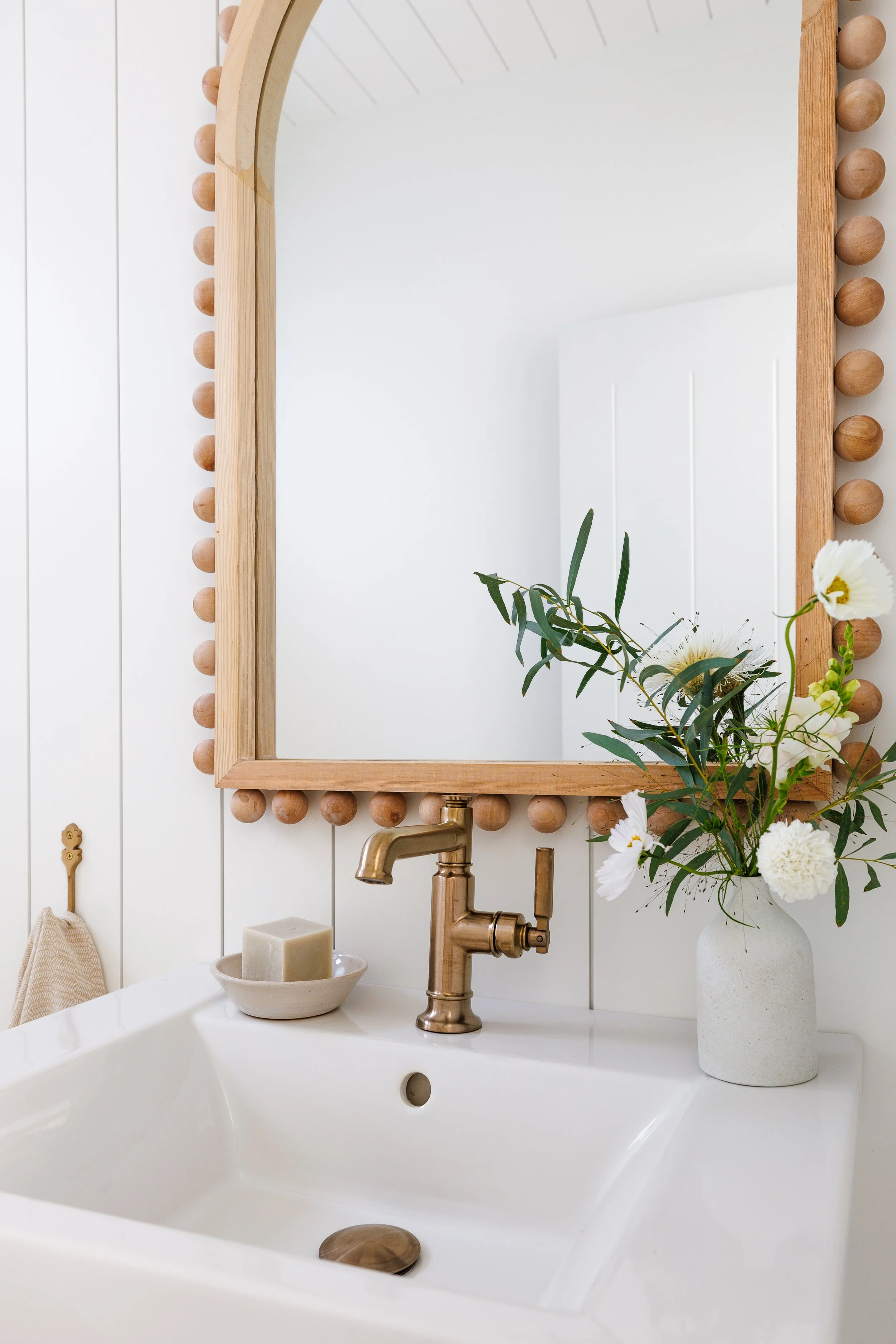 Close-up image of porcelain sink with spring flowers, gold faucet and arched wood mirror.
