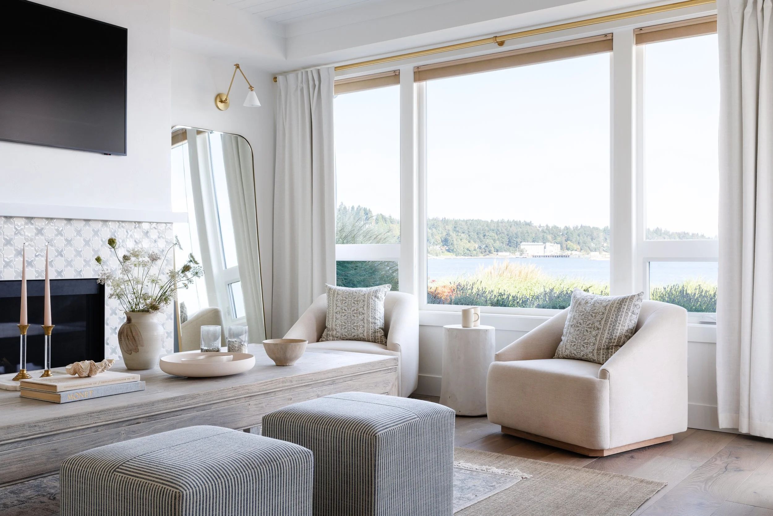 A bright living room with large windows overlooking a body of water and trees in the distance. The room features white sofas with patterned pillows, a rustic white coffee table with decorative bowls, a stack of books, and a vase with flowers. There is a wall-mounted flat-screen TV and a mirror leaning against the wall. White curtains frame the windows, and a gold wall sconce provides lighting.