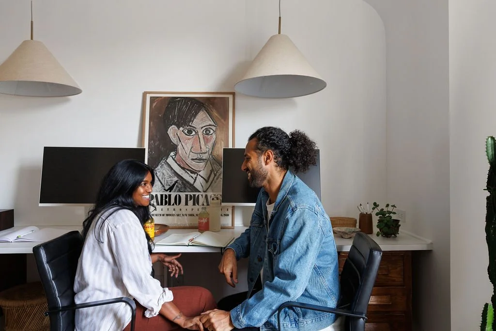 Syia and Rasila Herman discuss while holding hand at office desk.