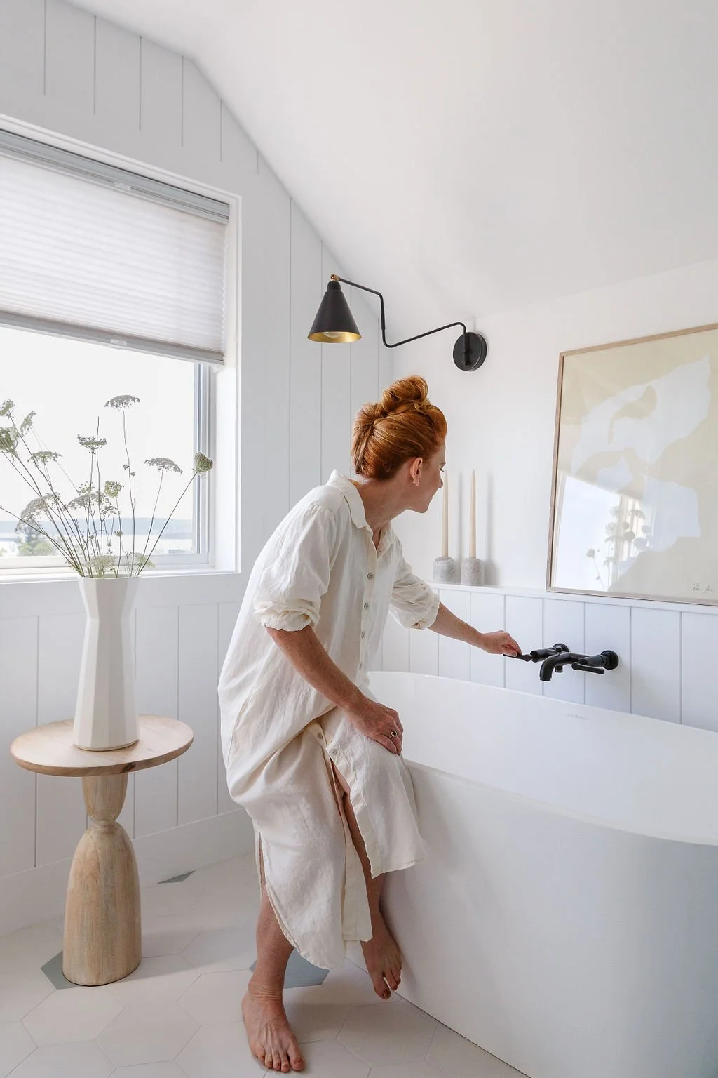 Woman with red hair sitting on edge of tub turning faucet on.