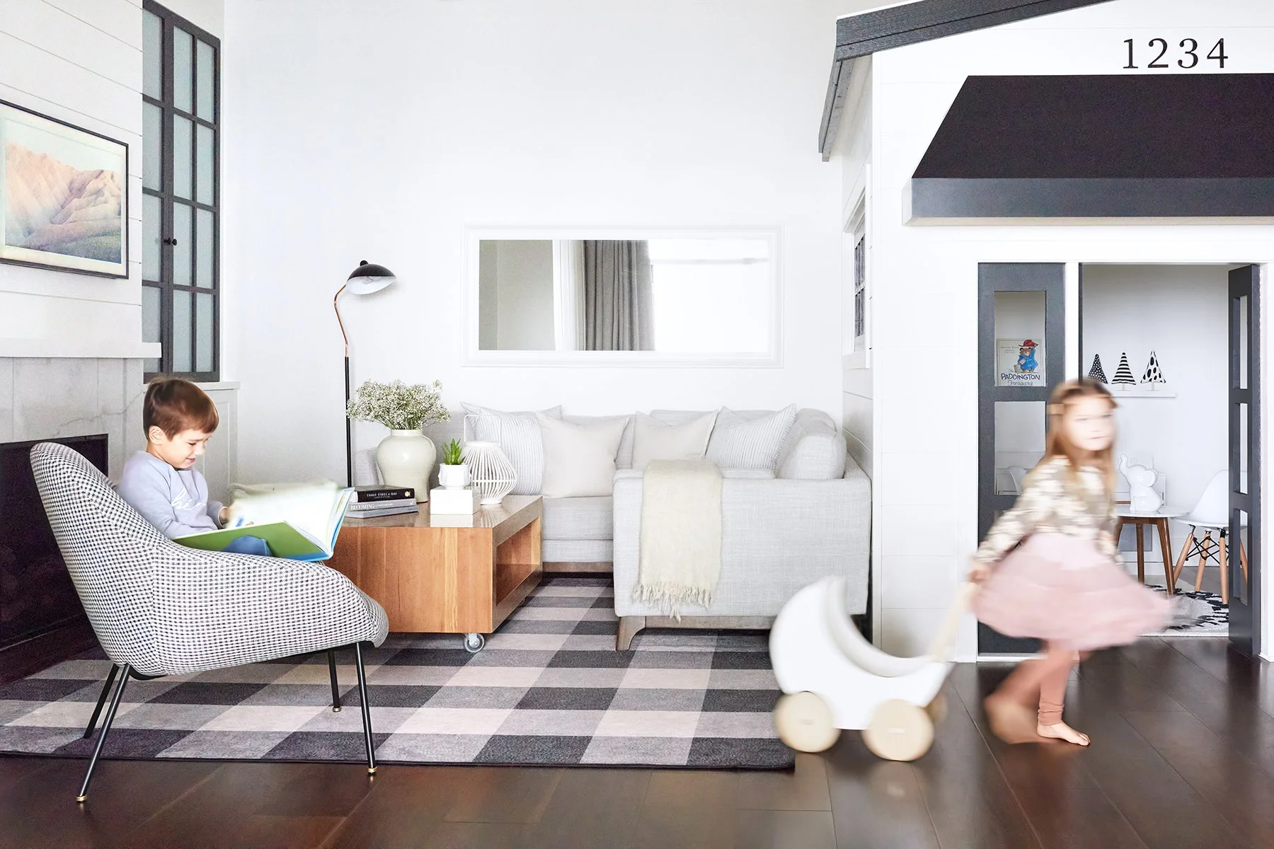 Photo of living room, boy sitting on chair reading a book and little girl pulling stroller in front of play house.