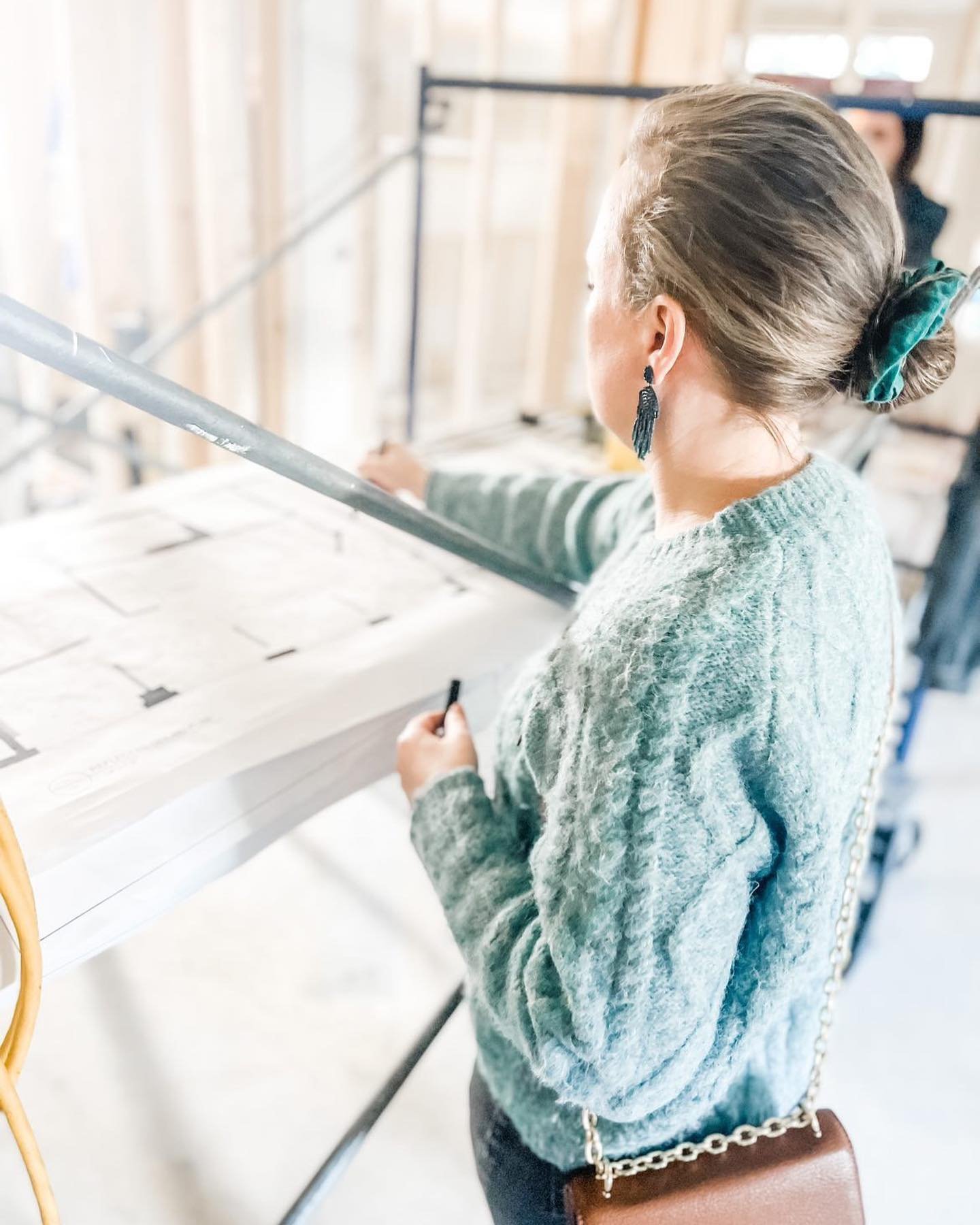 Interior Designer with hair tied with a green scrunchie and wearing a textured green sweater, holding a black pen, standing at a white table with plans , in a construction setting with wooden framing in the background.